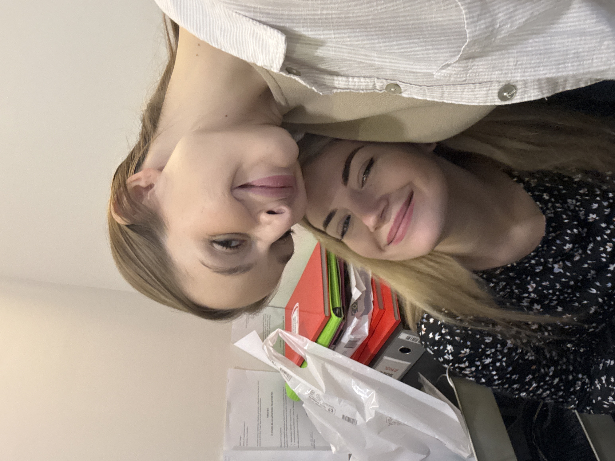 Two young adult females are depicted in a close-up, selfie-style photograph taken indoors. The woman in the upper frame, with light brown hair and a light-striped button-down shirt over a light undershirt, has her head tilted, resting on the head of the woman below her. She presents a subtle smile and looks directly at the camera. The woman in the lower frame, with blonde hair and a dark top featuring a light floral pattern, is smiling broadly with partially closed eyes, also looking at the camera. Both individuals have light complexions and dark eyebrows.
The background, visible to the left and behind the lower woman, reveals a cluttered indoor environment, likely an office or workspace. Visible objects include stacked documents and papers, a crumpled white plastic shopping bag, and a stack of ring binders and folders, notably a green one, several red ones, and a grey/black binder partially showing "210S". The wall in the background is plain and light-colored. The image itself contains no discernible features to confirm a specific location within the city of Budva, Montenegro; it depicts an interior setting.