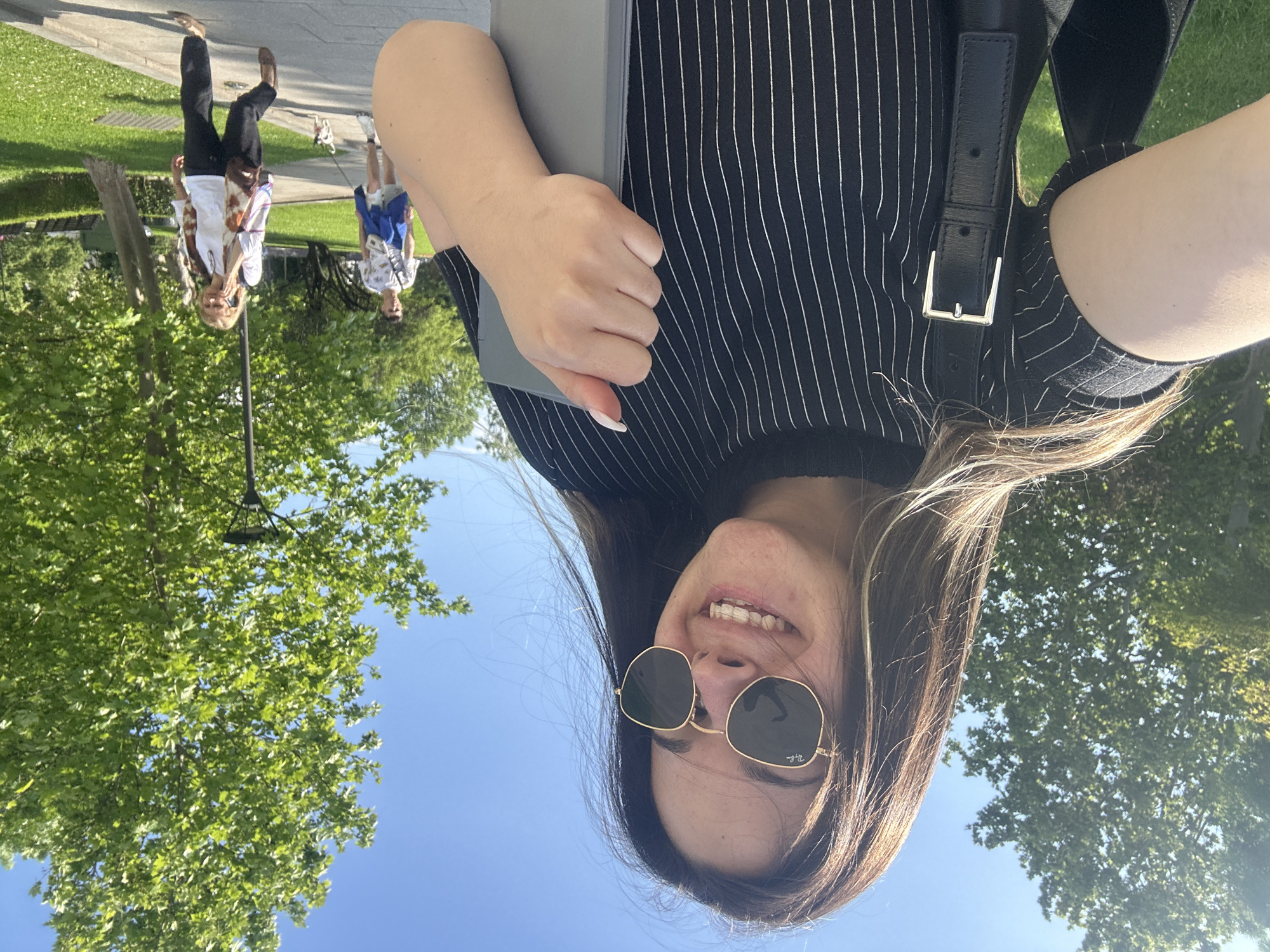 The image is a selfie taken by a woman with long brown hair, wearing sunglasses and a black and white striped top with a black strap bag. She is holding a gray object. In the background, there are green trees and a blue sky, suggesting an outdoor setting. Further in the background, two people are walking on a paved surface. The location appears to be a park or green space, possibly in Sofia, Bulgaria.