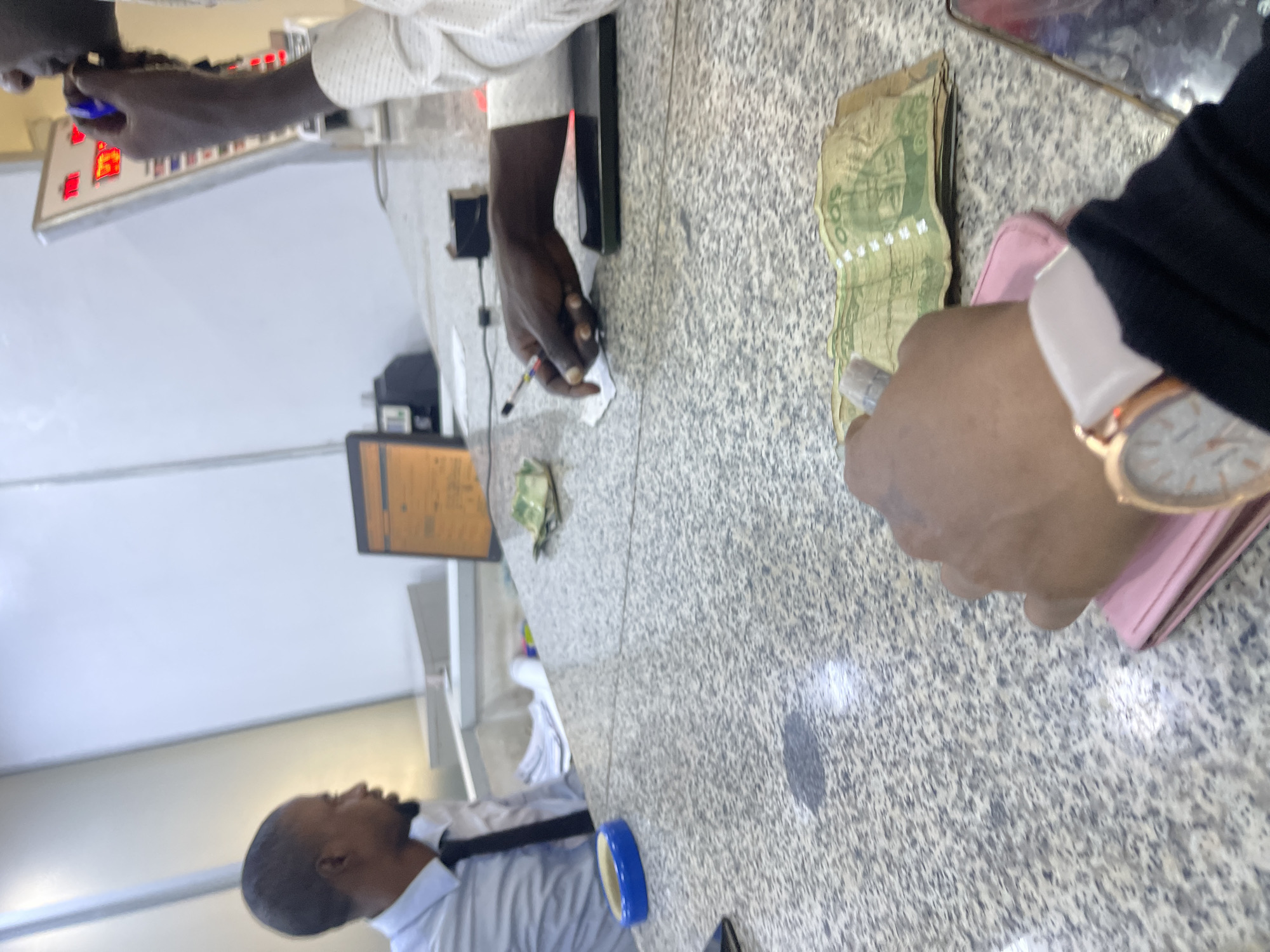 In a brightly lit room, two men and a woman are engaged in a transaction at a counter. The room is likely an exchange bureau or a bank in Jos, Nigeria, judging by the context of the currency.
On the left, a man in a light blue collared shirt and black tie faces away from the camera, seated behind a counter. His head is turned to the right, observing the activity. In front of him, a stack of papers is visible, suggesting administrative work. A blue round object, possibly an ashtray, sits on the counter near him.
In the center and right, a second man, partially visible, wears a white patterned shirt. He is leaning over the counter, holding a pen in his right hand, and appears to be writing on a small piece of paper. His left hand is visible near a smartphone lying flat on the counter. Scattered on the counter are several banknotes, predominantly green, suggesting Nigerian currency. One bundle of these notes is particularly visible in the foreground.
In the foreground, a woman's arm and hand are prominent. She is wearing a watch with a rose gold casing and a light-colored band. Her hand is reaching towards a pink wallet, which is positioned on the counter. The notes are spread out on the counter, some of them clearly visible with denominations printed on them, such as "500". The counter itself has a speckled granite-like surface.
Behind the man in the white shirt, a digital display board with red numerals and white lettering is mounted on the wall, displaying financial information. The overall environment suggests a busy financial transaction setting.