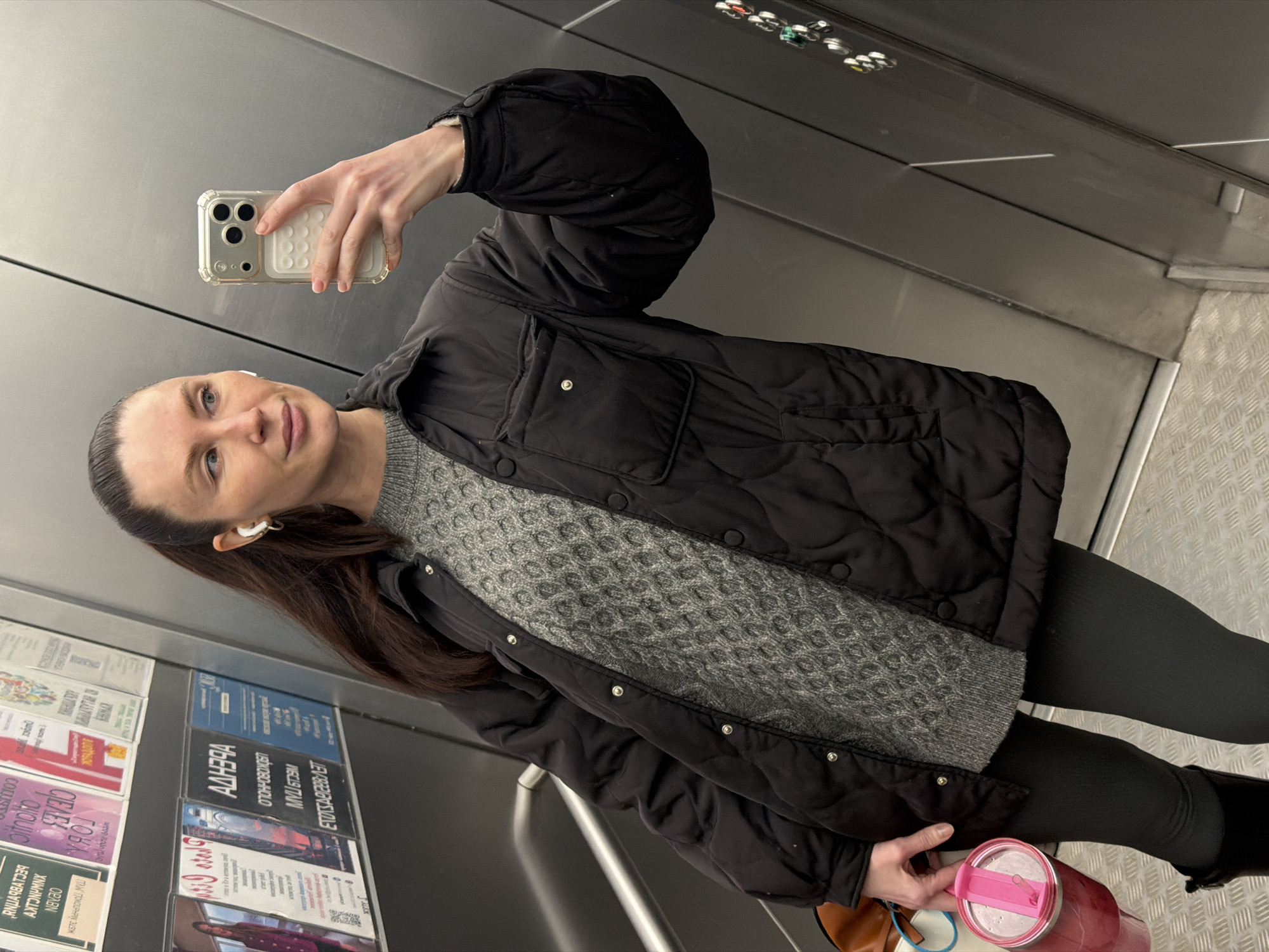 An adult woman is captured in a mirror selfie taken within what appears to be an elevator or similar enclosed public space. She holds an iPhone in a clear case with a white accessory for the selfie, angling it to capture her reflection. Her expression is neutral, and she wears white AirPods in both ears. Her dark hair is pulled back.
She is dressed in a black quilted jacket over a grey patterned knit sweater. She also wears dark leggings and dark boots. In her left hand, she holds a pink tumbler with a straw.
The background consists of metallic walls, with horizontal seams visible. The floor has a grey diamond plate pattern. On the left side of the image, display cases attached to the wall feature various posters or flyers, predominantly with Cyrillic text. These posters include titles like "АДЕНДА" (Agenda) on a blue poster, among other less discernible Russian text.
The setting, indicated by the Cyrillic text on the posters and the metallic, utilitarian decor, is consistent with the provided context of Perm, Russia. The image does not provide cues for the time of day or specific weather.