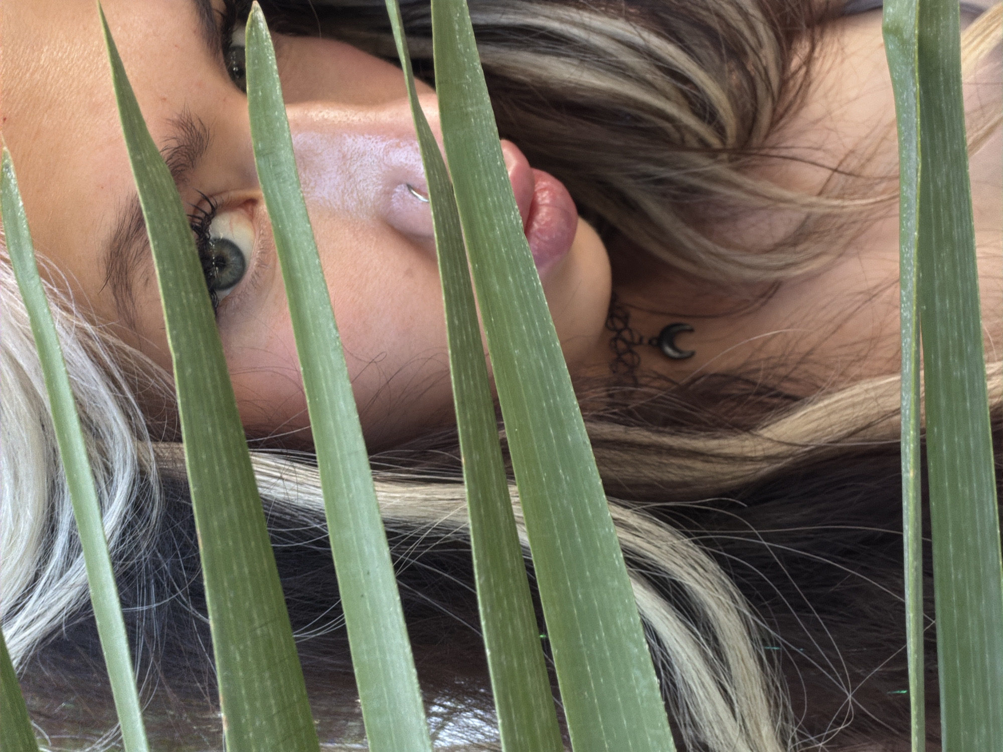 A close-up, eye-level shot depicts a person's face, partially obscured by green, slender leaves. The person is lying down, with their head tilted to the side, showing an eye, part of the face including a nostril and lip, and hair. The eye is grey-green. The hair is dark with streaks of blonde. A silver nose ring is visible in one nostril. A crescent moon-shaped pendant hangs from a thin necklace around the neck. The leaves create a layered foreground, drawing attention to the partial view of the person's face. The setting's location in Bar, Montenegro is not discernible from the image's content.