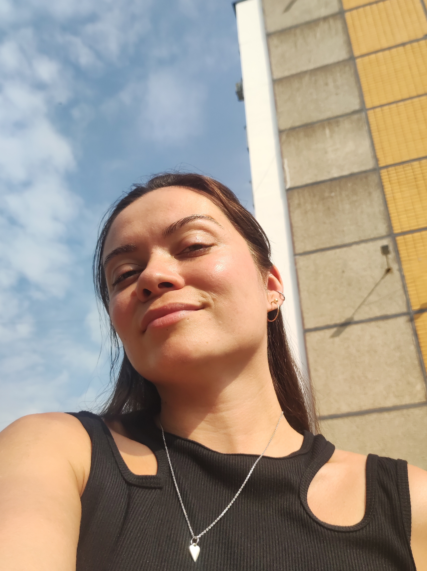 A low-angle, close-up selfie shows a young woman with brown hair and brown eyes, wearing a black sleeveless top and a silver heart pendant necklace. She is outdoors, with a blue sky and clouds above her. To her right, a portion of a building is visible, appearing to be made of concrete and yellow tiles. The overall lighting suggests it is daytime, likely late afternoon due to the direction of the sun casting shadows. The woman has a slight smile and her face is illuminated by the sun, highlighting her cheekbones. Her right ear has two small hoop earrings. The background suggests an urban environment, possibly Chaikovsky, Russia, given the architectural style of the building.