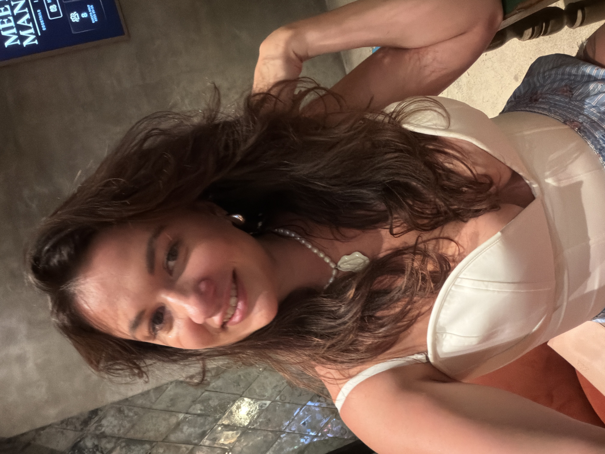 A woman with long, dark brown hair is captured in a selfie at a dimly lit venue in North Kuta, Indonesia. She is wearing a white, strapless corset-style top and a pearl necklace with a large pendant. The background features a textured wall with reflective surfaces on the left and a digital display on the right showing "MEE MAN" and other text. The woman is smiling and looking at the camera, with her right arm resting on her head and her left hand touching her hair. The overall atmosphere suggests a casual, nighttime setting.