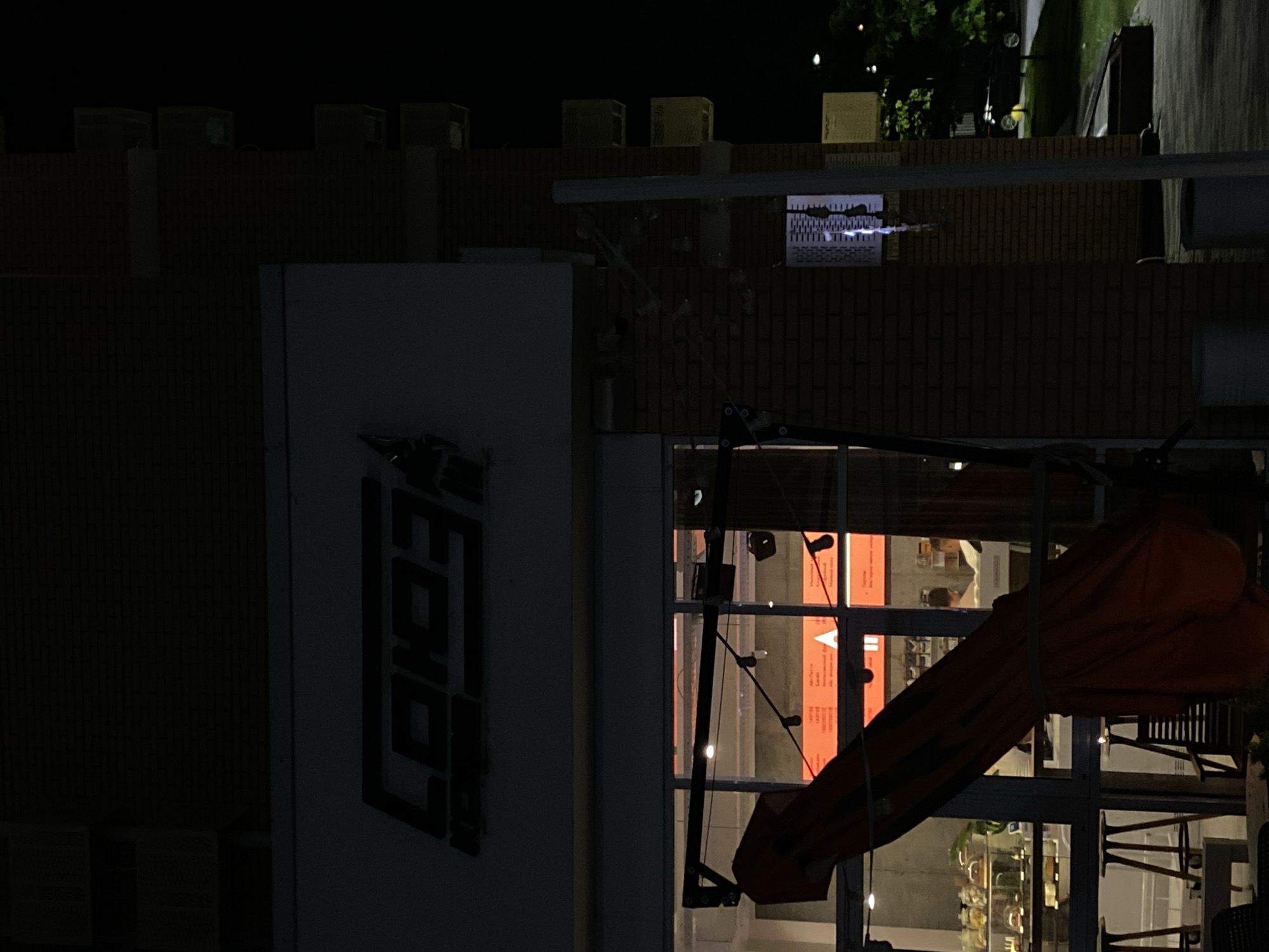 The image shows a night view of a building with the logo "COOO3" visible on its facade. The building appears to be a cafe or restaurant, with large glass windows revealing the interior. Inside, a counter with display cases of food items and people can be seen. An orange umbrella is tilted in the foreground, partially obscuring the view. The building is made of brick and is located in the city of Vishnyaki, Russia. The scene is dimly lit, with streetlights and interior lights providing illumination.
