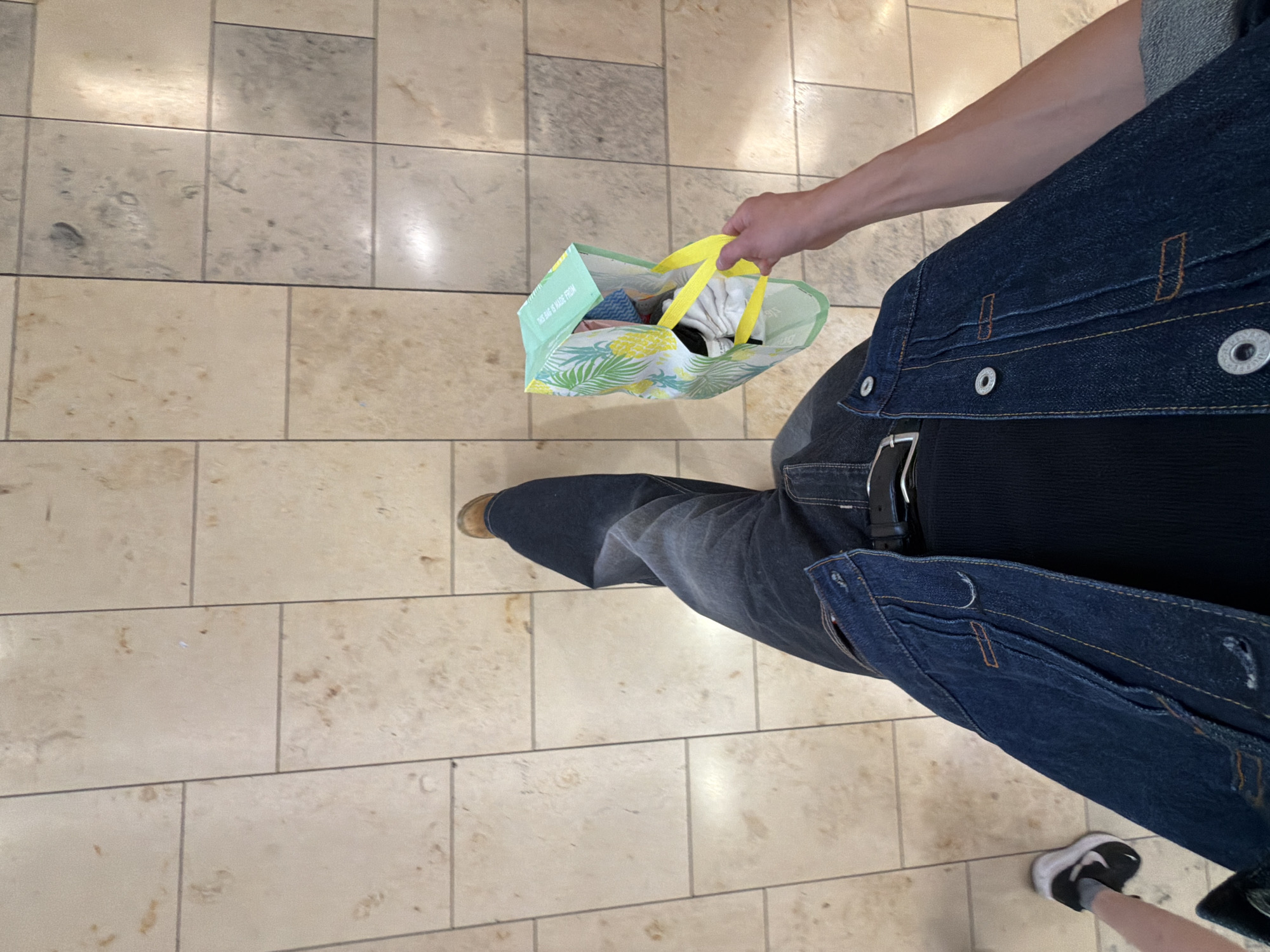 The image shows a person's legs and torso from a high-angle perspective, standing on a tiled floor. The person is wearing dark wash jeans, a dark ribbed tank top, and a dark denim jacket. They are holding a reusable shopping bag with a tropical print, containing some items. To the left, another person's legs and feet are visible, wearing sneakers and grey socks. The tiled floor is light beige with darker grout lines and some subtle discoloration. The scene appears to be indoors, possibly in a commercial setting like a shopping mall or a store. The lighting suggests a well-lit indoor environment. There is no specific indication of the location being in London, United Kingdom.