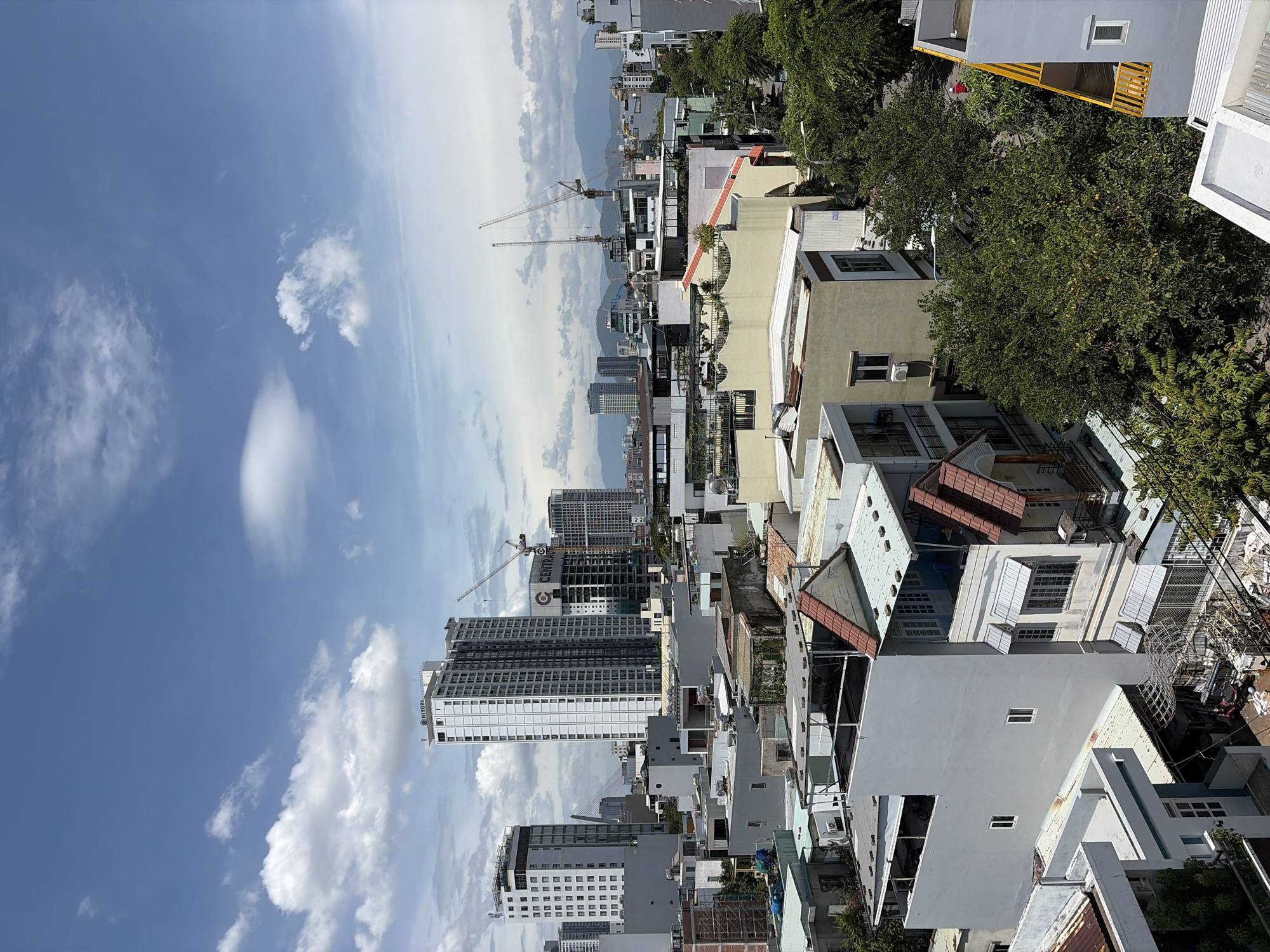 The image depicts a panoramic view of a densely populated urban area in Da Nang, Vietnam, under a partly cloudy sky. The scene is dominated by a mix of modern high-rise buildings and lower-rise residential structures, characteristic of a developing city. In the distance, a range of mountains can be seen, suggesting the city's geographical proximity to a natural landscape.
Several construction cranes are visible, indicating ongoing development and infrastructure projects within the city. The architecture of the buildings varies, from sleek, contemporary skyscrapers with glass facades to more traditional, multi-story houses with tiled roofs and balconies. Many of the residential buildings appear to be built close together, with some showing signs of wear and tear, such as rusted metalwork and faded paint.
Greenery is present in the form of trees and rooftop gardens, offering a contrast to the concrete and steel structures. Streets are visible winding through the city, with parked vehicles and some scattered pedestrians. The lighting suggests it is daytime, with sunlight casting shadows on the buildings and ground.
Specific identifiable elements include a tall, white building with the name "HIYORI" visible at the top. Another prominent building features a large "G CENTRA" sign. The overall impression is one of a bustling city undergoing rapid growth and modernization, nestled between natural scenery.