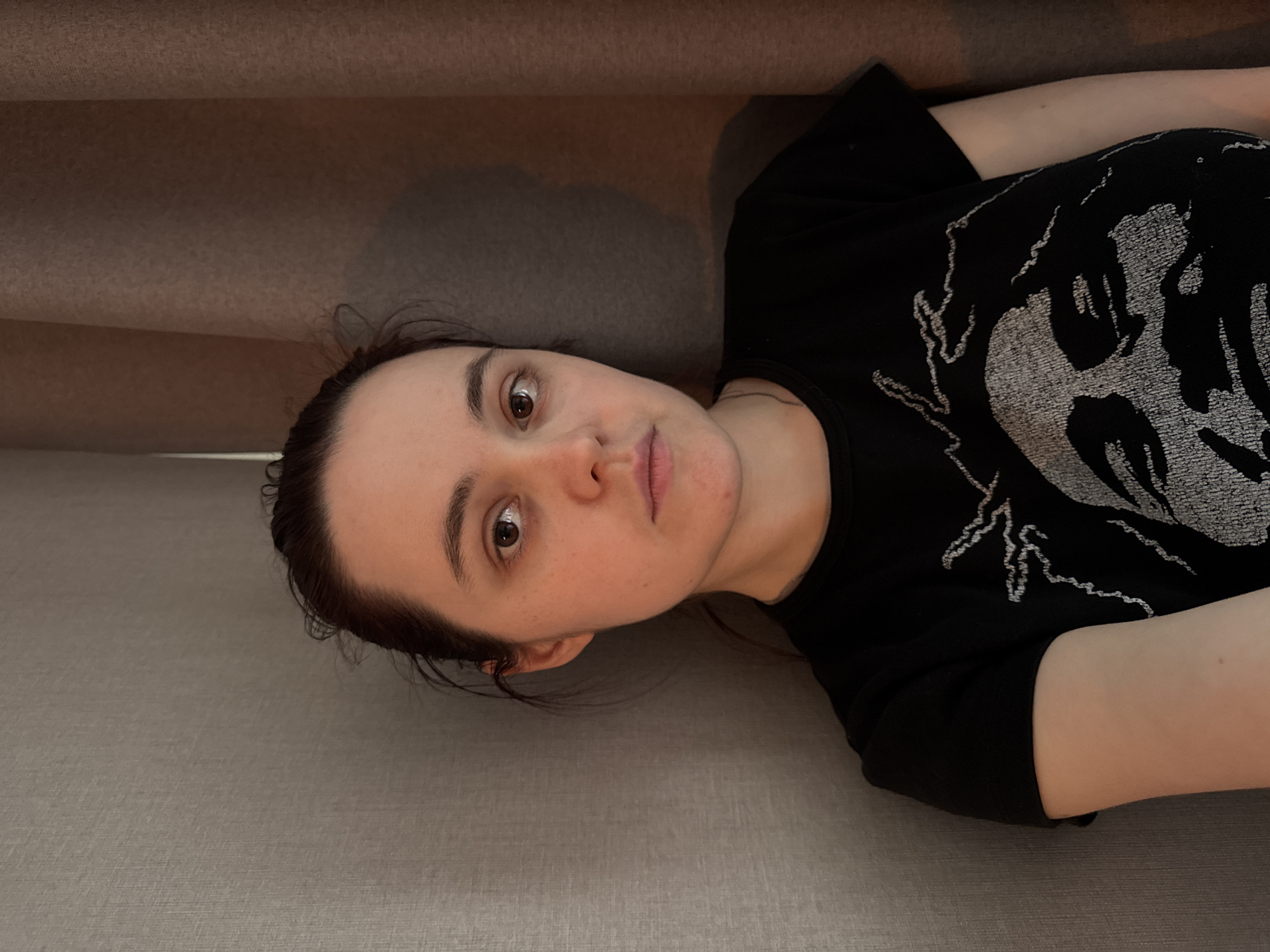 A fair-skinned woman with dark hair pulled back is lying on a light grey surface, possibly a couch or bed. She is wearing a black t-shirt with a large, light-grey skull graphic on the front. The image is taken from an overhead perspective, slightly angled. A shadow falls from an unseen object above her, possibly a shelf or ledge. There are no indications of the location being in Izhevsk, Russia.