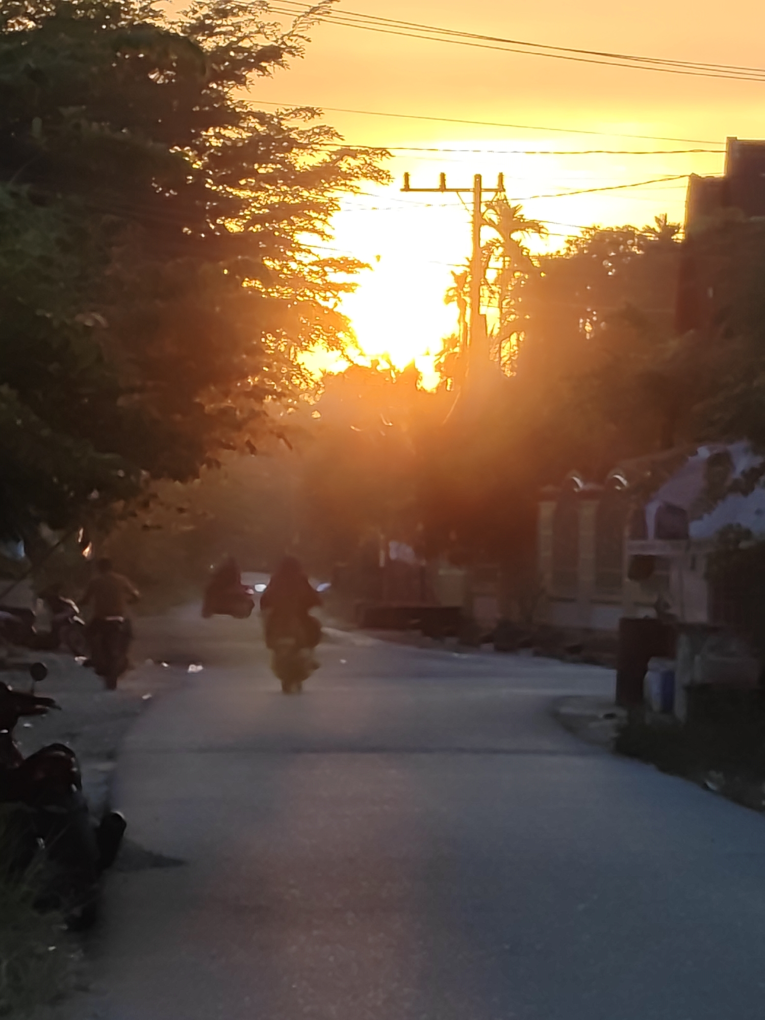 The image depicts a street scene at sunset in Lhokkulam, Indonesia. The sun is low on the horizon, casting a bright orange and yellow glow across the sky and silhouetting trees and buildings. Several people are riding motorcycles on the paved road. In the foreground, a motorcycle is parked on the left side of the road. The street is lined with trees and some buildings, with utility poles and wires visible in the background. The overall atmosphere is hazy due to the low sun angle.