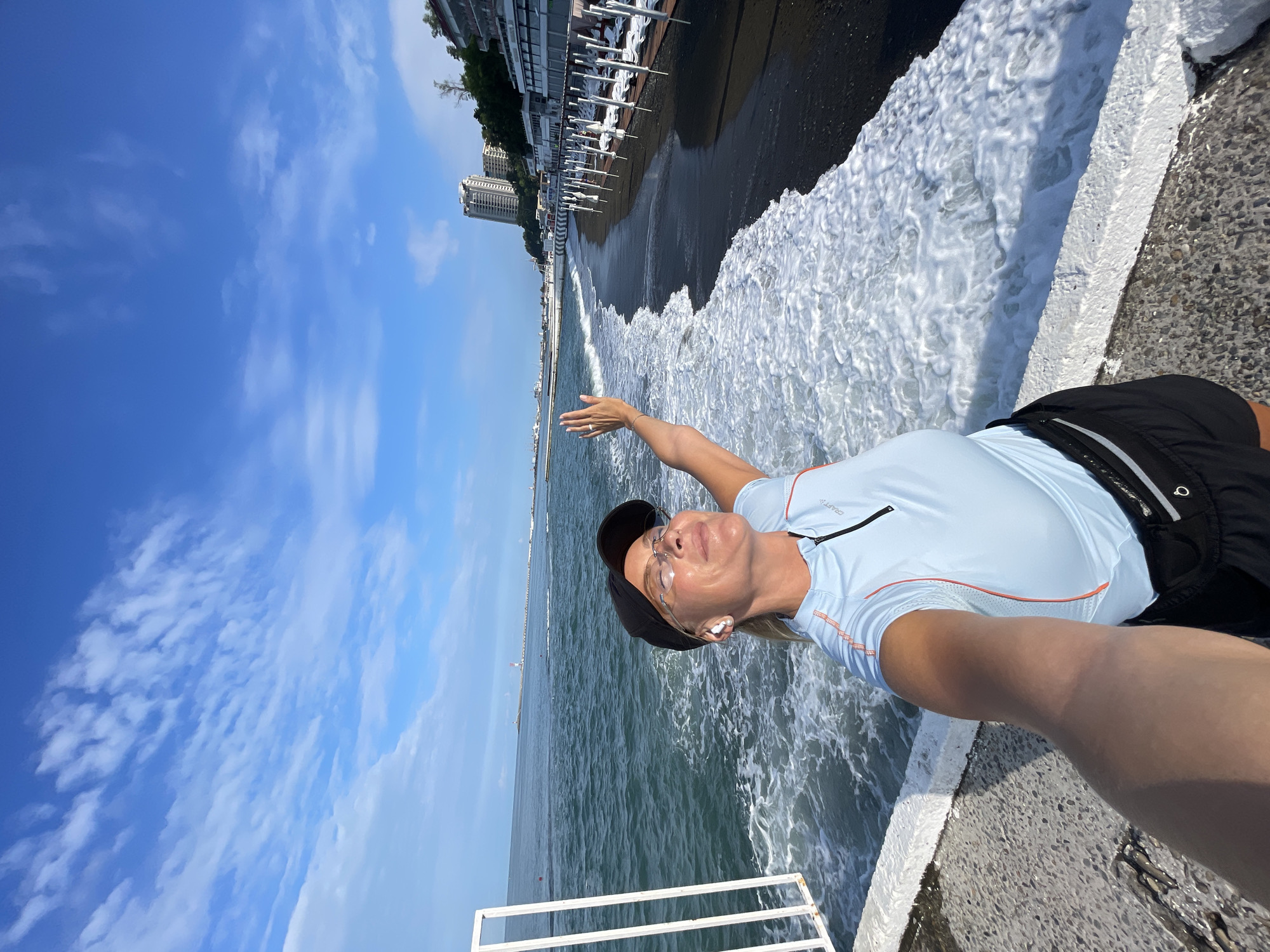 A woman in athletic wear is taking a selfie on a sunny day at the beach in Sochi, Russia. She is wearing a light blue athletic shirt, black shorts, a black baseball cap, and glasses. She has on wireless earbuds. The woman is smiling with her eyes closed and her right arm is raised, with her palm facing forward. She is standing on a concrete walkway that borders a black sand beach. The sea is to her left, with waves breaking on the shore and creating white foam. In the background, there are several buildings, including a tall, modern high-rise, as well as beach chairs and umbrellas set up on the sand. The sky is blue with scattered white clouds. The scene suggests a moment of enjoyment and relaxation by the sea.