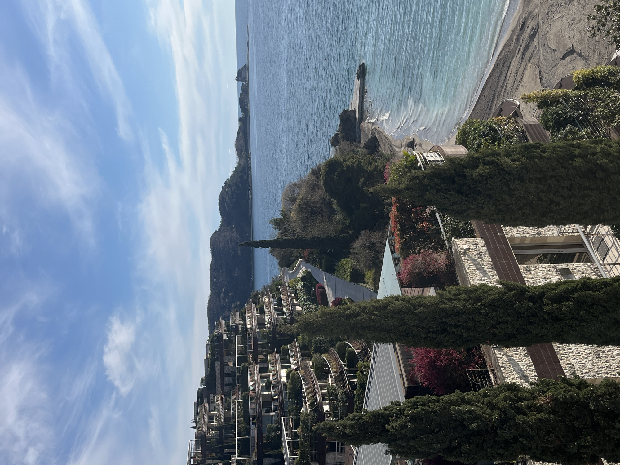 The image depicts a coastal landscape, likely in Budva, Montenegro. The scene is captured from an elevated vantage point, revealing a series of modern terraced buildings nestled into the hillside. These buildings feature balconies with rounded trellis-like structures. Tall, slender cypress trees are interspersed among the buildings.
The coastline consists of a strip of dark sand or shingle beach, adjacent to a calm, blue sea. In the distance, a land formation juts out into the water. The sky is mostly clear with scattered clouds.