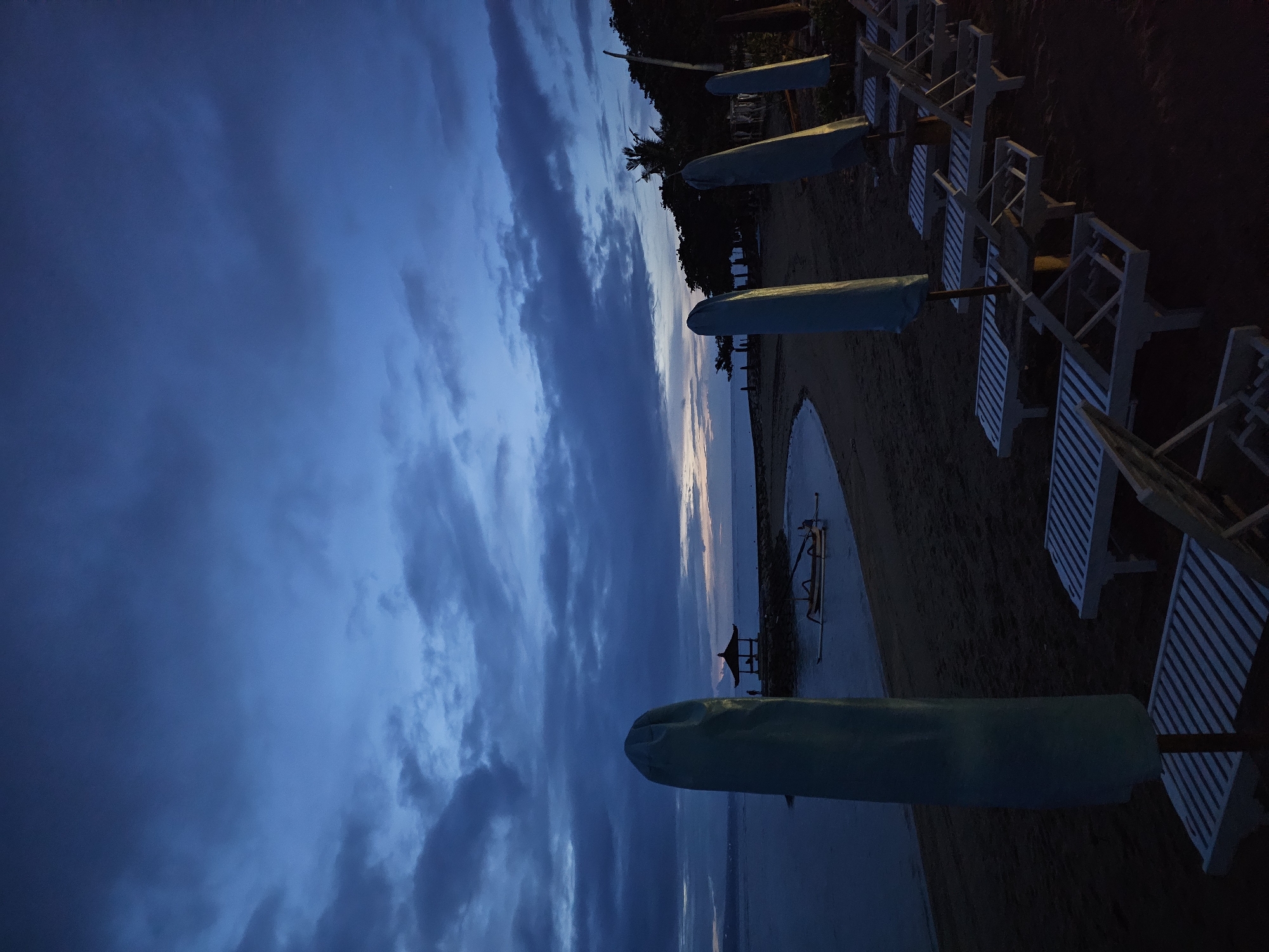 The image depicts a beach scene at dusk in Denpasar, Indonesia. The sky is filled with dark, heavy clouds, hinting at either an approaching storm or a recent rainfall. A sliver of light from the setting sun can be seen on the horizon. The beach itself is dark and partially obscured, with rows of covered beach umbrellas and unoccupied white beach chairs. A small structure, possibly a gazebo, is visible in the distance near the water's edge. The overall atmosphere is serene and quiet, suggesting the end of the day at the beach.