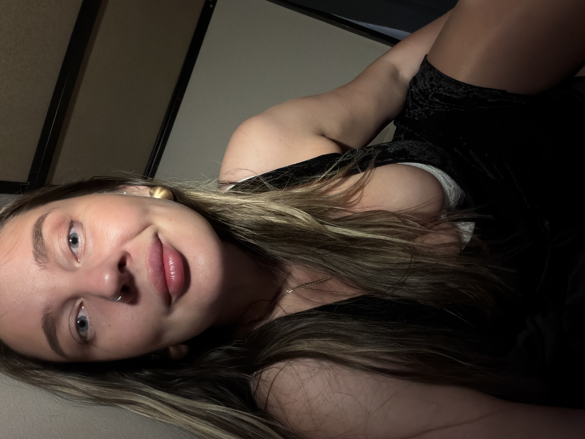 A young woman with fair skin, light eyebrows, and blue eyes, is lying down in a dimly lit room. She is wearing a black velvet dress with a white bra visible. She has long, brown and blonde hair and gold earrings. A thin gold necklace is around her neck, and a ring pierces her nose. The background features a wall and a dark frame. The location is not discernible.