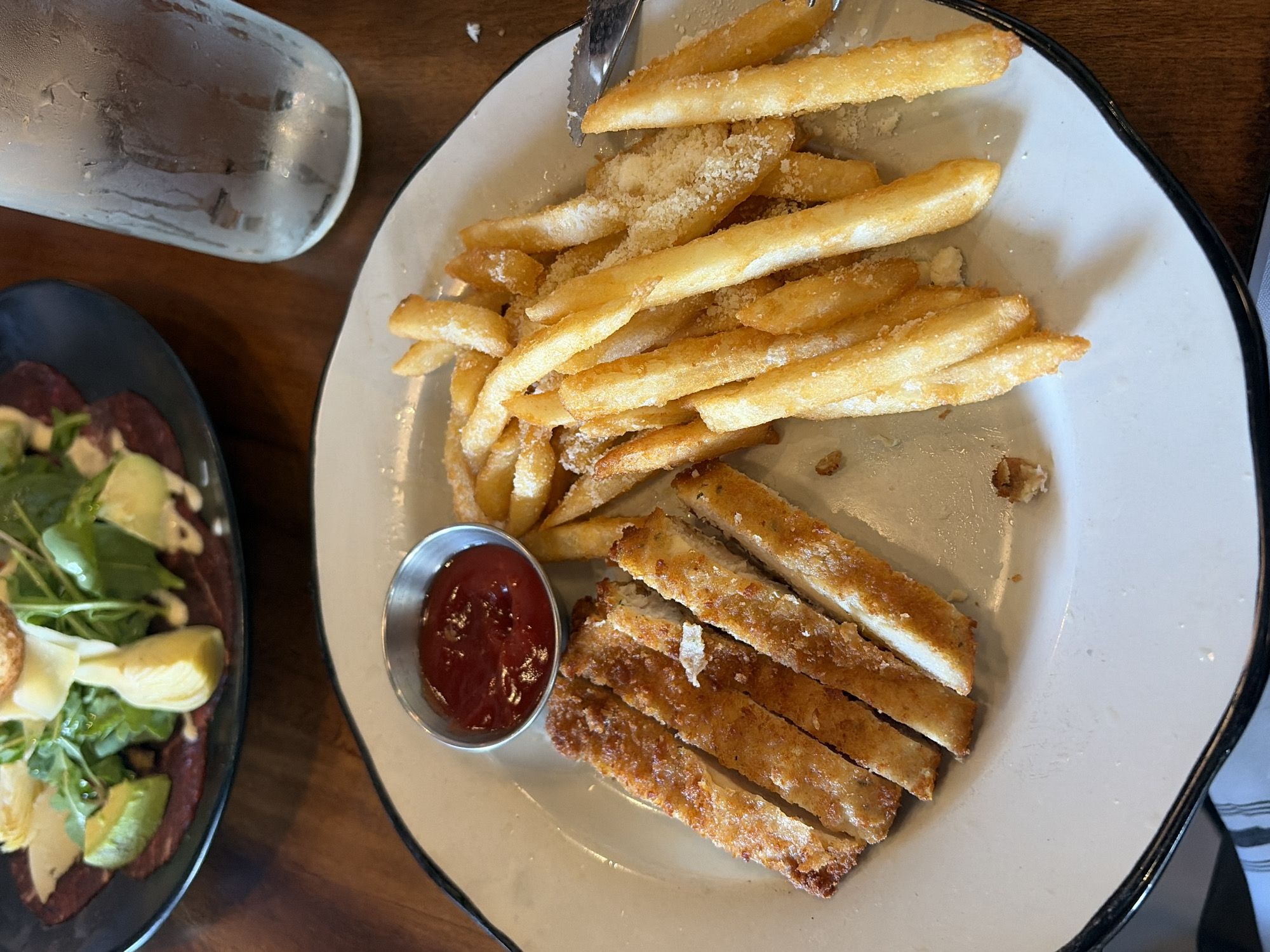 A table in Key Biscayne, United States of America, is set with a glass of water and two plates of food. One plate holds a salad with greens, avocado, and other ingredients arranged on a dark base. The other plate has a serving of french fries with parmesan cheese, alongside strips of breaded protein and a small metal container filled with red sauce. A fork rests on top of the fries.