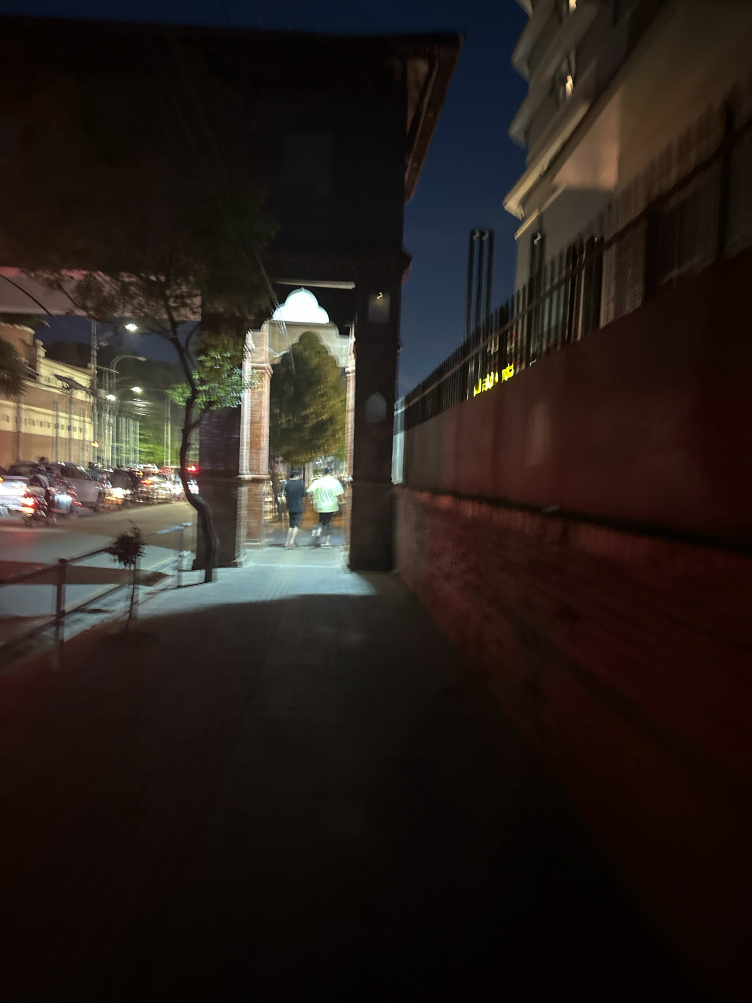 An outdoor, street-level, night time photograph captures a pedestrian walkway and street scene in Kathmandu Metropolitan City, Nepal. The perspective is from a sidewalk looking towards a street with traffic and buildings on either side. A dark, overhanging structure is in the foreground on the left, casting a shadow over the walkway. On the right side of the walkway, a red brick wall runs along the edge, with a metal fence above it. Beyond the fence, a multi-story building with lit windows is visible.
Further down the walkway, a large, arched gateway made of brick is present, framing two individuals walking away from the camera. One person is wearing a light-colored t-shirt and shorts, and the other is in a dark t-shirt and shorts. The street to the left is filled with vehicles, appearing blurred due to motion and low light conditions, with their headlights and taillights creating streaks of light. Streetlights illuminate the scene, casting a glow on the pavement and surrounding structures. Trees are visible, with one in the middle ground appearing as a dark silhouette. The sky above is a dark blue, indicating nighttime. The overall atmosphere is one of a busy urban environment at night.