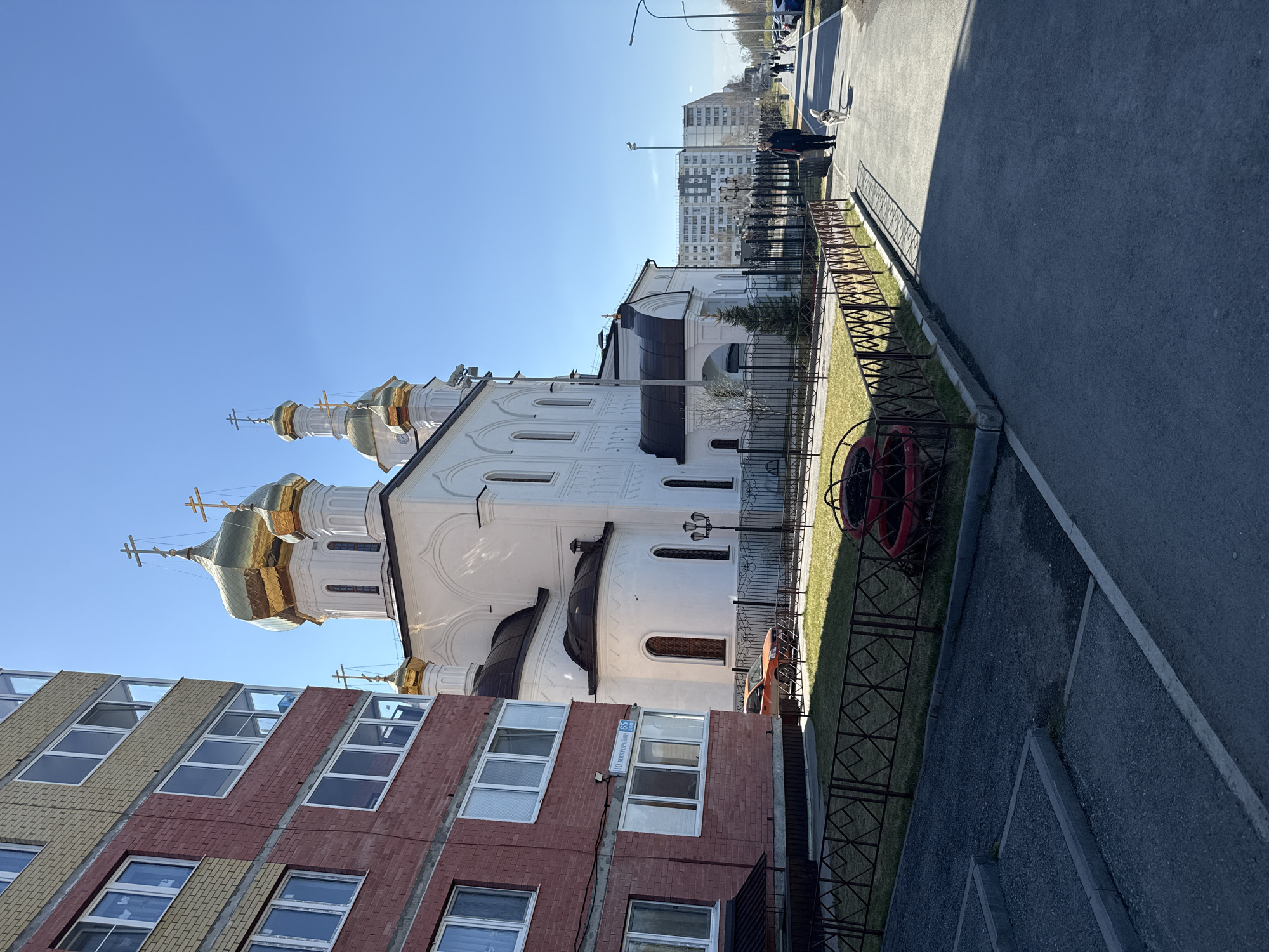 The image shows a white-walled building with gold domes, likely an Orthodox church, juxtaposed with a multi-story brick building with multiple windows. The buildings are located in a city setting, possibly Tobolsk, Russia. A black metal fence separates a grassy area from a paved road. A person and a dog are walking on the sidewalk. There are other buildings visible in the background. The sky is clear and blue. A sign on the brick building says "10 ЮНОСТИ, 15".