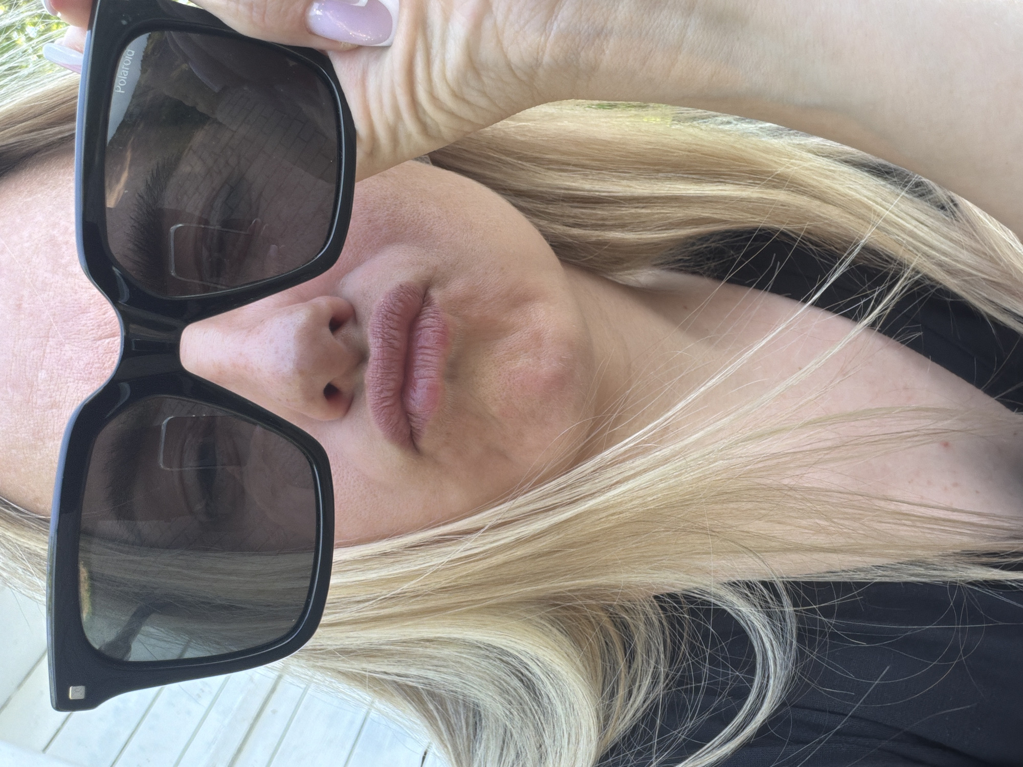 A woman is holding a pair of black Polaroid sunglasses up to her face. Her blonde hair is visible, as is part of her black top. Her manicured hand is holding the sunglasses. The location is not identifiable as Votkinsk, Russia from the provided image.