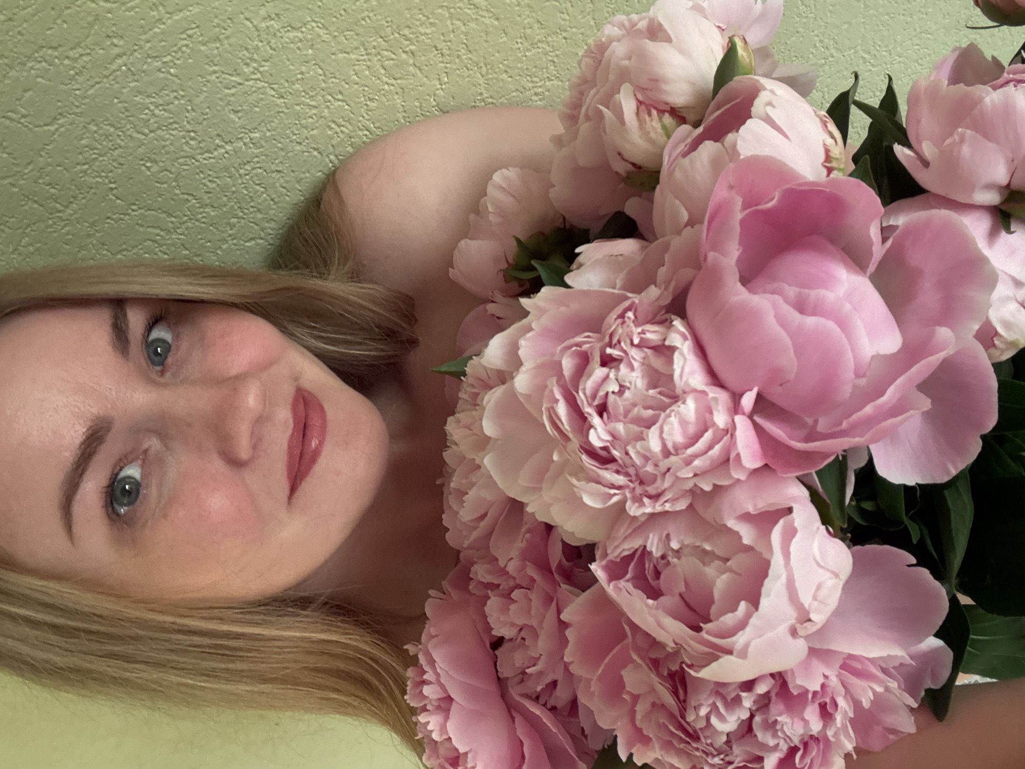 A blonde woman with blue eyes and light makeup holds a large bouquet of pink peonies in front of a textured pale green wall. She is looking at the camera and smiling slightly. Her shoulders and upper chest are visible. The image appears to be taken indoors. There is no information to indicate the location in Chaikovsky, Russia.