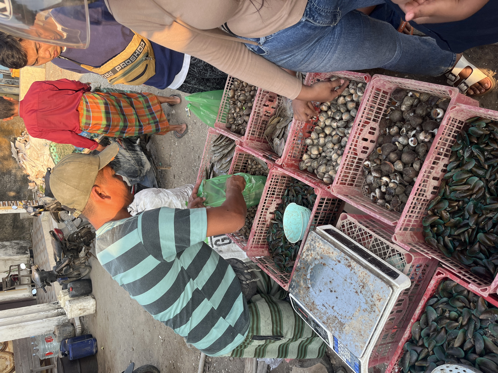 A man in a striped t-shirt and shorts is selling seafood in a market in Kedungmalang, Indonesia. He is standing next to a pile of baskets filled with mussels and clams. Another man in a red shirt and plaid sarong is standing behind him, and a third man in a blue shirt and yellow fanny pack is to his right. A woman in a tan shirt and jeans is reaching into a basket of clams. In the background, there are motorcycles and water containers. The ground is dusty and uneven.