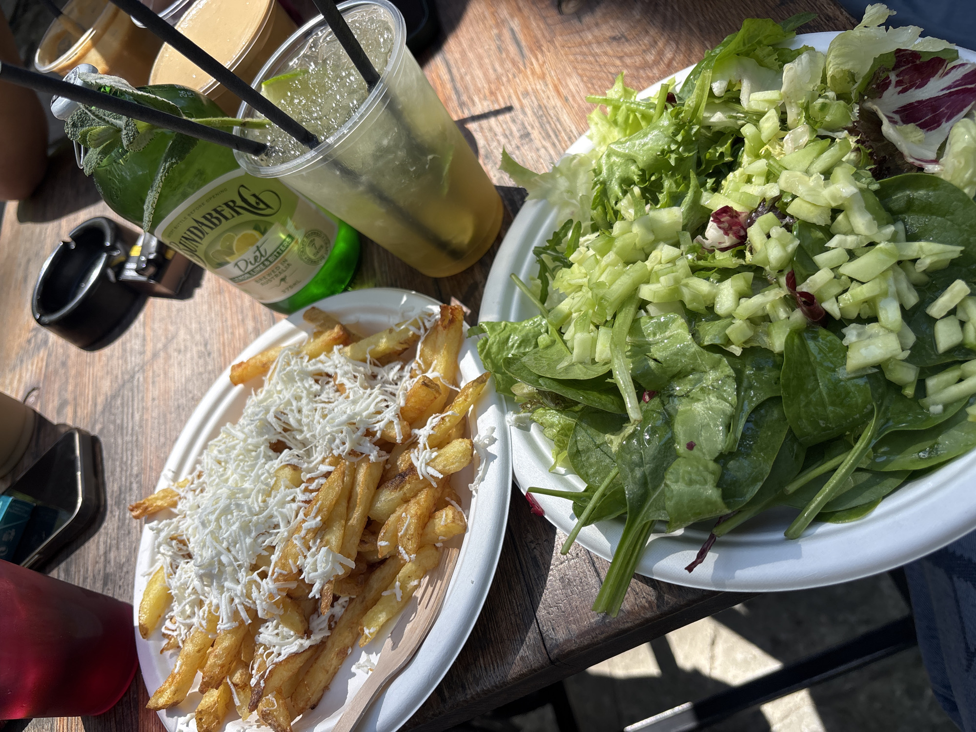 The scene is an outdoor dining setting, presumably at a restaurant or cafe. On a wooden table are two white plates. One plate is filled with french fries generously topped with shredded white cheese. The other plate contains a salad of mixed greens, including spinach, lettuce, and what appears to be diced cucumber. Next to the plates is a bottle of Bundaberg Diet Lemon Lime & Bitters and a clear plastic cup with a light-colored drink and ice. Black straws are in both the cup and the Bundaberg bottle. An ashtray is visible near the bottle. The lighting suggests a sunny day. The image was taken in Sofia, Bulgaria.