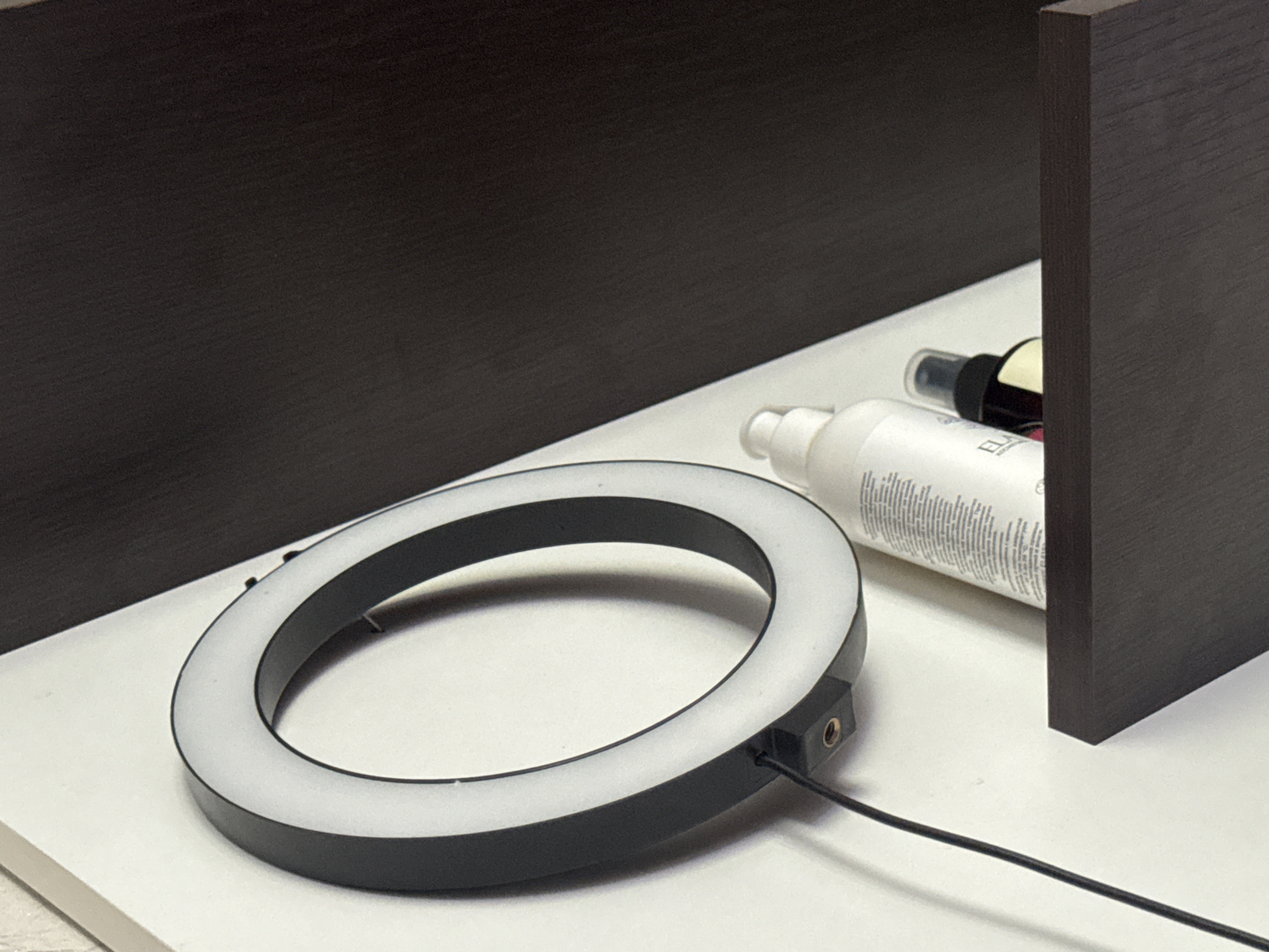 The image shows a close-up of a ring light and two bottles on a white surface, possibly a desk or shelf. The ring light, with its black casing and illuminated white interior, is the most prominent object in the foreground. A black cable is connected to the side of the ring light and extends out of the frame. Behind the ring light, two bottles are visible. The one on the left is a white spray bottle with text printed on it. The bottle on the right is darker, possibly a cosmetic or skincare product. The background consists of dark, textured surfaces, likely shelves or cabinet doors, suggesting an indoor setting. One of these dark surfaces is positioned vertically on the right side of the frame, partially obscuring the bottles. The lighting in the image is soft and diffused, possibly from the ring light itself. There are no people or animals in the image, and no clear indication of the time of day or weather conditions. The overall impression is that of a product setup, possibly for photography or videography, or simply a collection of personal care items arranged on a surface. The visible text on the white bottle is illegible due to its small size and the resolution of the image.