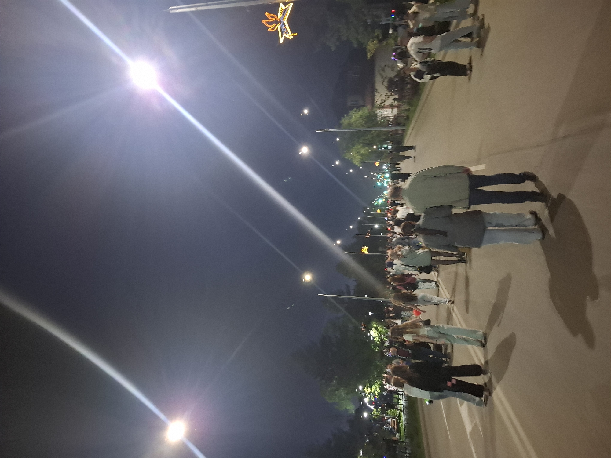The image depicts a nighttime scene on a street in Svobodny, Russia, likely during a public event or festival. A large crowd of people is walking along a paved road, illuminated by streetlights and bright overhead lights. The sky is dark, indicating it is late evening or night. On the right side of the road, there is a decorated streetlamp with a star-shaped illuminated ornament. Trees and buildings are visible on the sides of the street, some with illuminated windows. The overall atmosphere appears to be lively and social, with people casually dressed and interacting.