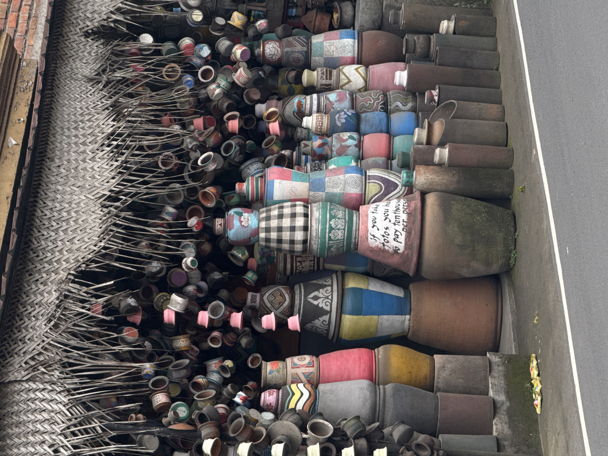 This image captures an outdoor display of numerous handcrafted ceramic pots and vases in what appears to be a market or artisan workshop in Ubud District, Indonesia. The scene is densely packed with items, showcasing a wide variety of shapes, sizes, and decorative styles.
On the left side, hundreds of smaller, often unpainted or lightly decorated terracotta pots are arranged on what looks like a drying rack or display, with thin wooden or bamboo sticks holding them. Behind this, a large, intricately woven mat-like structure, possibly made of natural fibers, serves as a backdrop or part of a shelter.
Towards the center and right, larger and more elaborately decorated pots are stacked vertically and horizontally, creating colorful columns. These pots feature diverse patterns, including geometric designs, floral motifs, checkerboard patterns, and abstract swirls, in a palette of blues, greens, reds, yellows, and earthy tones. Some are plain brown or grey. The arrangement suggests an extensive collection, possibly for sale or artistic display.
No people are visible in the photograph, but the organized display implies human activity related to crafting and commerce. The setting appears to be outdoors during the daytime, possibly under an overcast sky or shaded area, as the lighting is diffused with no harsh shadows.
A notable detail is handwritten text visible on a large pinkish-brown pot in the lower right quadrant. It clearly reads: "If you take fotos you have to pay ten thousand Rpt/person," indicating a charge for photography at this location, likely in Indonesian Rupiah. The far right of the image shows a paved surface, likely a road or sidewalk, bordered by a concrete curb with some small weeds and litter.