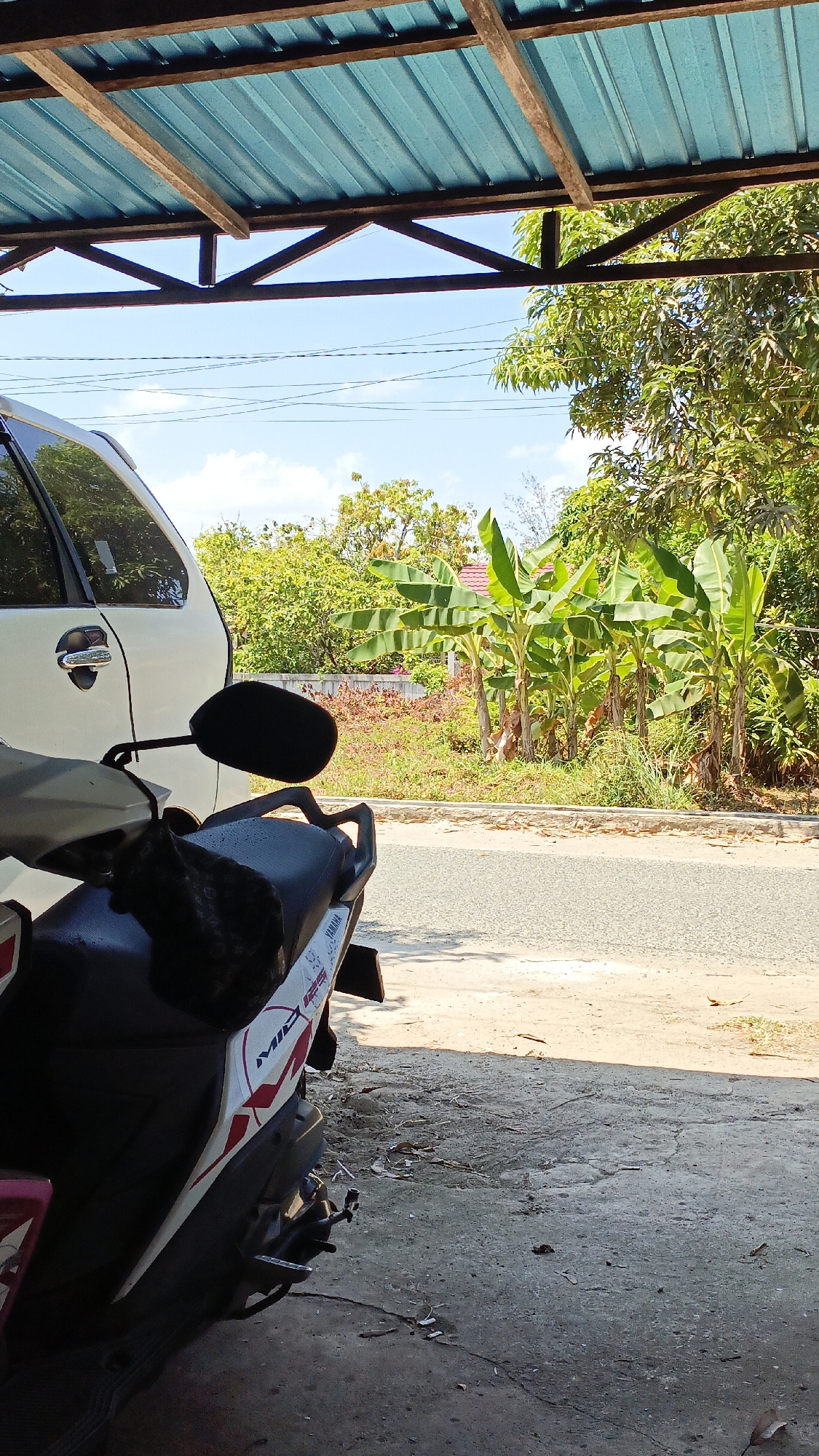 In Gusunge, Indonesia, this image captures a view from under a weathered blue corrugated metal roof supported by wooden beams. A white car is parked to the left, its door handle visible. In the foreground, the seat and part of the body of a white motorcycle with red and black decals, including the branding "MID 125" and "YAMAHA", are visible. A dark cloth object rests on the motorcycle seat. Beyond the parked vehicles, a paved road runs horizontally. The background features a row of banana trees with large green leaves, and further back, lush green foliage and trees line the horizon under a bright blue sky with scattered white clouds. A small red-roofed building is partially visible amongst the trees. The ground in the foreground is a mix of cracked concrete and loose dirt.