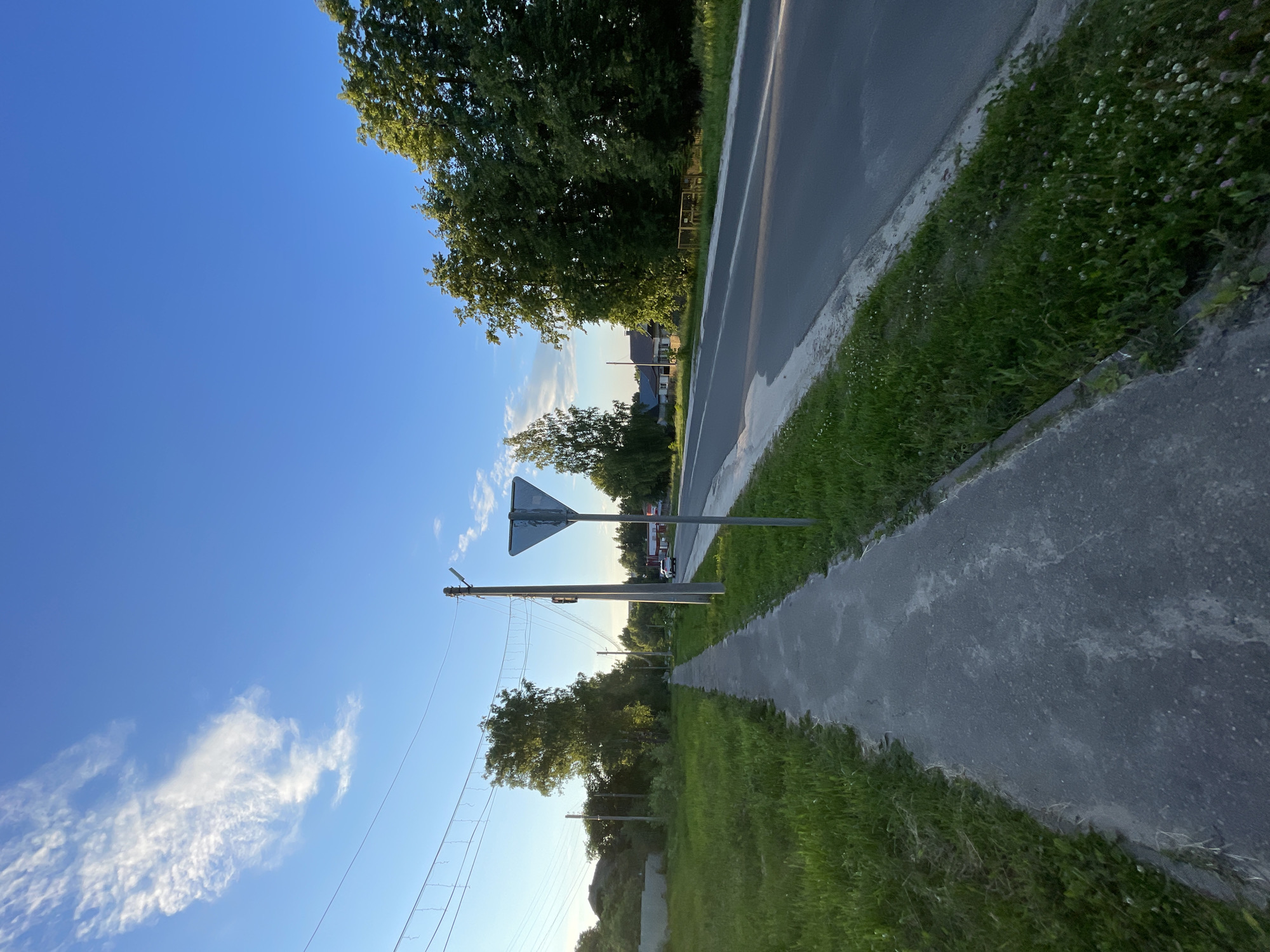 The image depicts a quiet, tree-lined road in the village of Krasnye Baki, Russia, during what appears to be late afternoon or early evening. A paved sidewalk runs along the left side of the road, bordered by lush green grass. The road itself is asphalt and appears to be in good condition, with a white line marking the edge.
On the right side of the road, a traffic sign, a blue and white inverted triangle, is prominently displayed on a metal pole. Beyond the sign, a white car is visible driving away from the camera. Further down the road, houses with red roofs can be seen nestled among the trees. The trees are predominantly deciduous, with dense green foliage, and some of them are illuminated by the warm light of the setting sun. The sky is a clear, vibrant blue with a few wispy white clouds. Overhead, power lines are strung between utility poles, with decorative string lights also visible. The overall impression is one of a peaceful rural setting on a pleasant day.