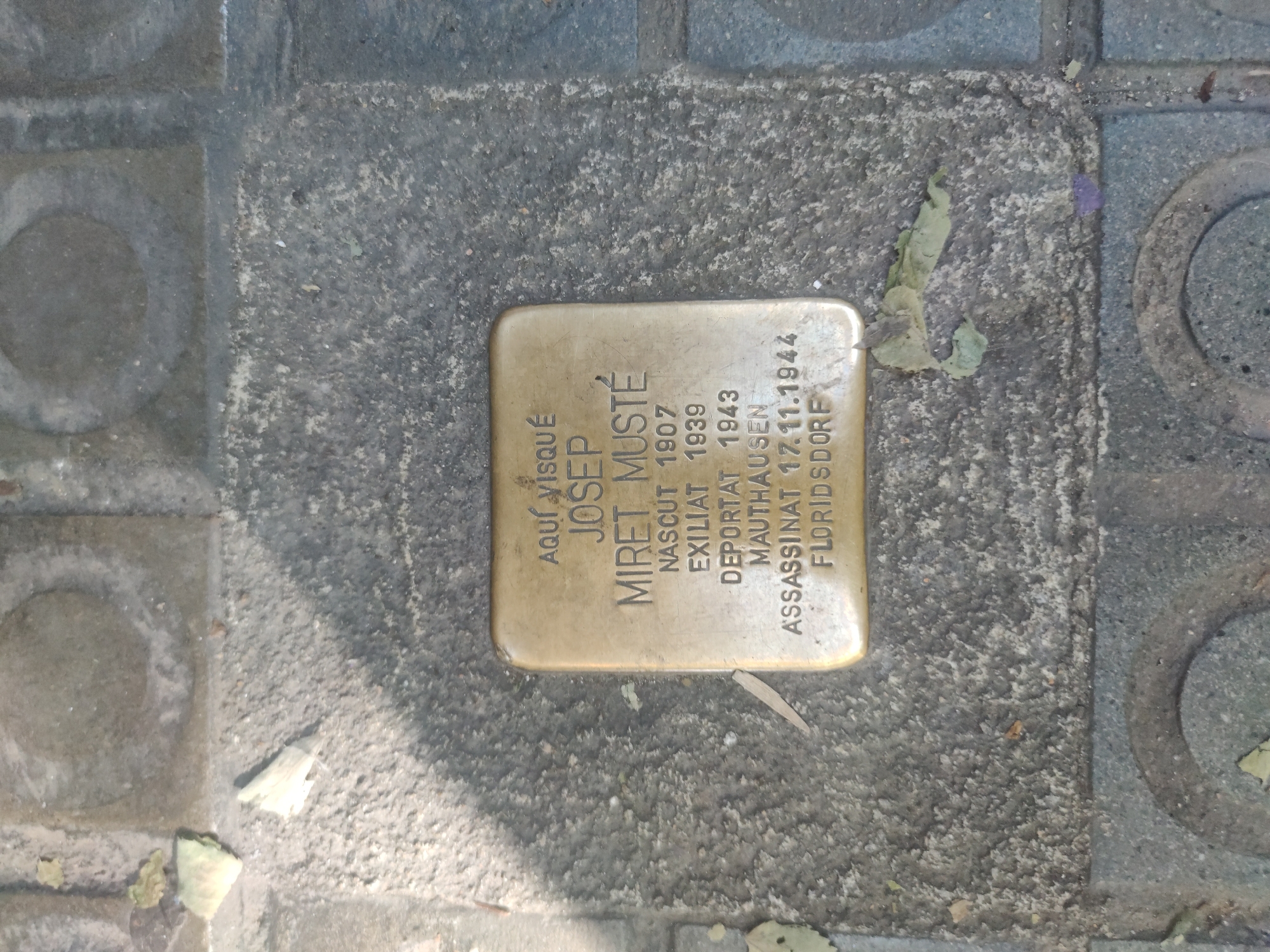 The image shows a "Stolpersteine" or "Stumbling Stone" set into the pavement. The brass plaque commemorates Josep Miret Musté, born in 1907, exiled in 1939, deported in 1943 to Mauthausen concentration camp, and assassinated in Floridsdorf on November 17, 1944. The phrase "AQUÍ VISQUÉ" (Here lived) in Catalan is inscribed at the top left of the stone. The stone is surrounded by grey paving stones with worn textured surfaces. The location is likely in Barcelona, Spain, given the Catalan language on the stone.