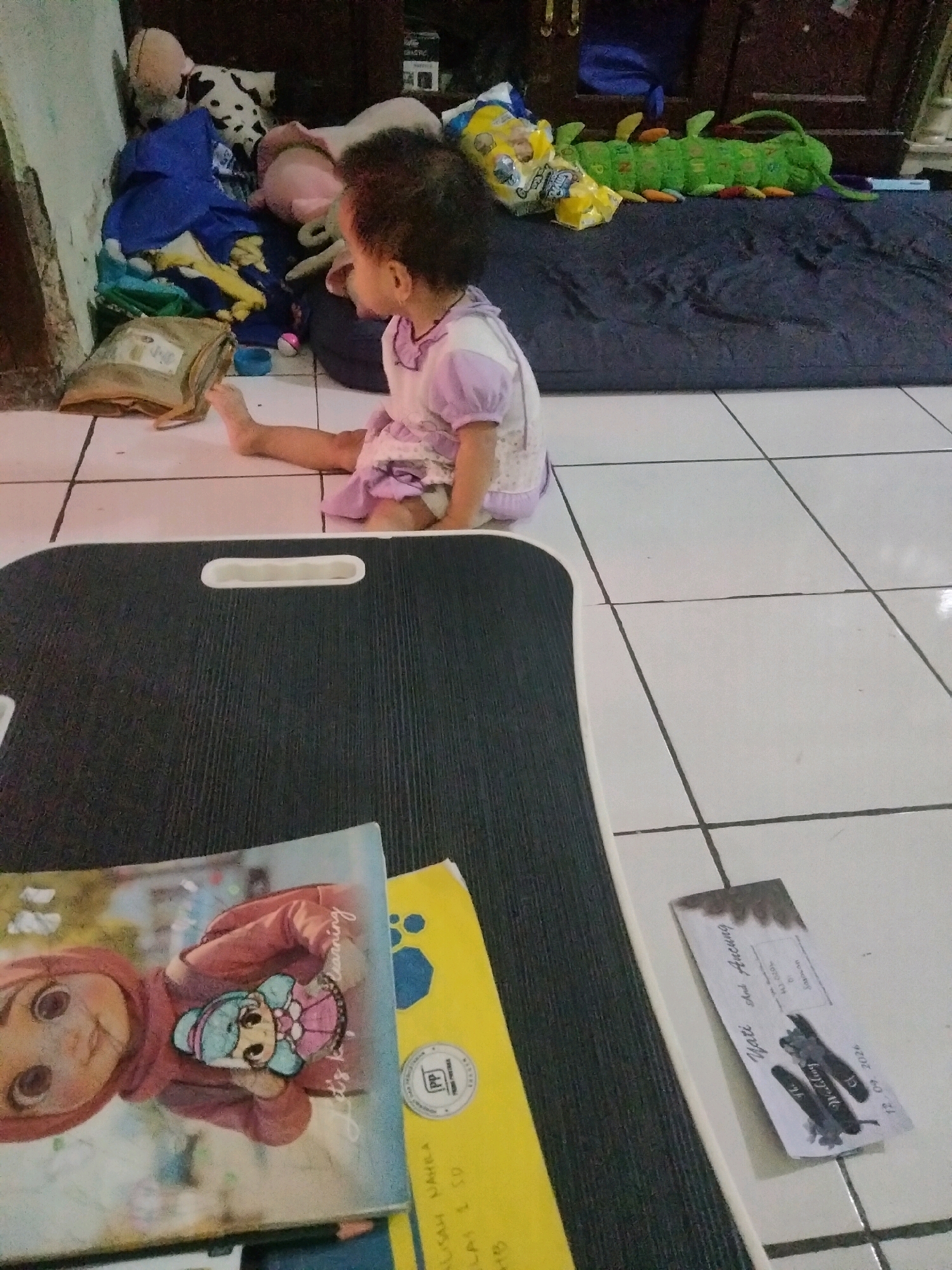 A young child with dark, curly hair, wearing a light purple and white dress, sits on a white tiled floor in what appears to be a residential setting in Suka Sari, Indonesia. The child is positioned facing away from the camera, looking towards a pile of toys and blankets. To the child's left, a brown paper package rests on the floor. In the foreground, a black tray with a white handle is placed on the tiles, and on top of it are two books, one with a colorful illustration of a doll-like character and text that includes "Let's Go Learning," and another yellow book with some writing. A small, dark object, possibly a ticket or a folded piece of paper, lies on the floor to the right of the tray. The background features a wall with a wooden cabinet and various items, including stuffed animals and a long, green caterpillar-shaped toy. The overall scene suggests a child's play area or a living space.