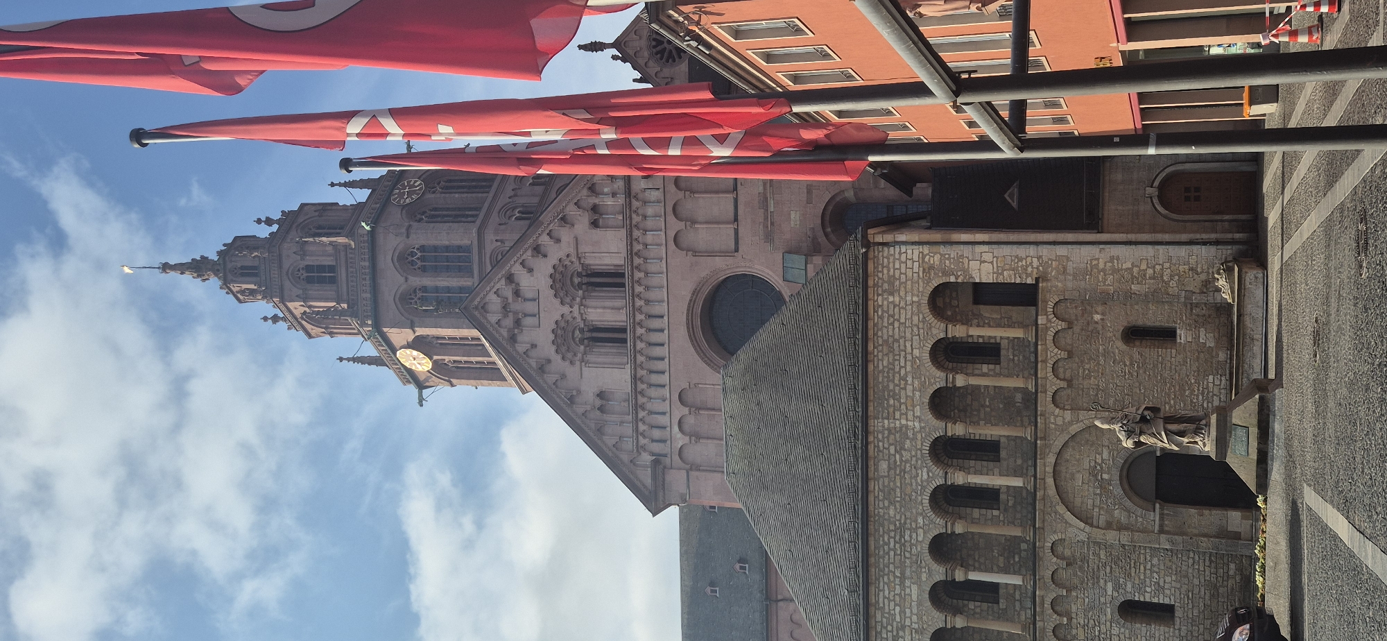 The image is a vertically oriented photograph of the Mainz Cathedral in Mainz, Germany. Red banners with white symbols are draped across the top of the frame. The cathedral's facade is made of pink sandstone, with intricate architectural details including arched windows and decorative carvings. A large clock face is visible on one of the towers. In the foreground, to the right, is a stone statue of a robed figure holding a staff, standing in front of a stone building with arched doorways and small windows. The ground is paved with cobblestones and has a patterned design. The sky above is blue with scattered white clouds. The scene suggests a sunny day, possibly in the late morning or afternoon given the light. There is no text visible in the image.