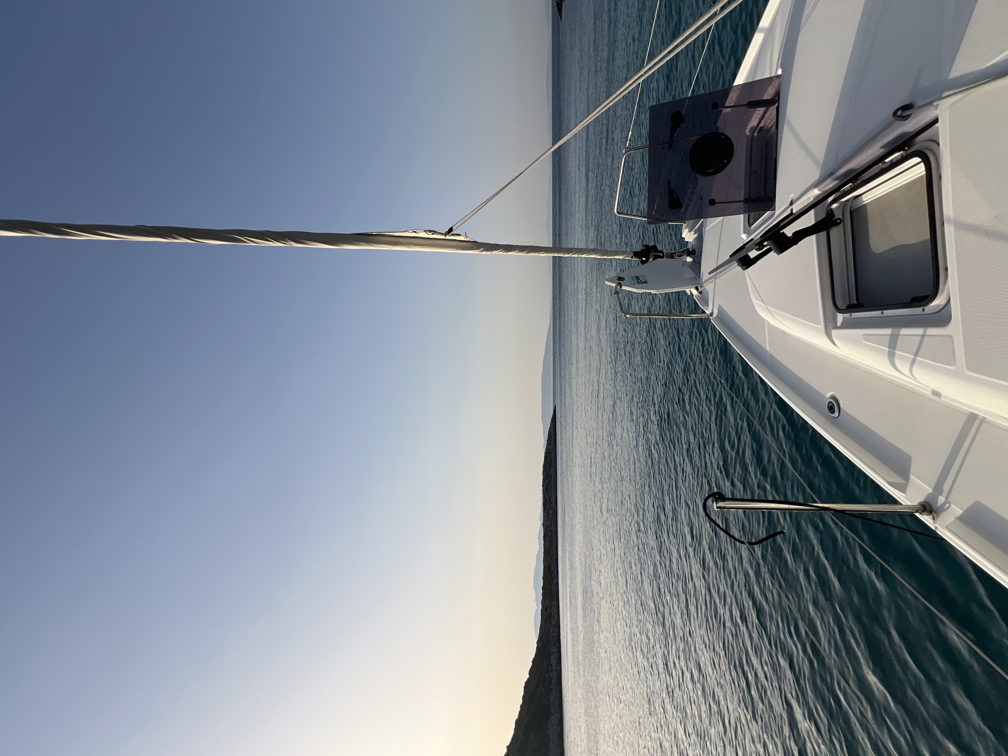An outdoor, eye-level, point-of-view shot shows the front of a white sailboat navigating calm, blue-green water under a clear, pale blue sky. The sun, low on the horizon, casts a soft light on the scene, creating a gentle glow. In the distance, a hazy, dark green landmass with a rocky shoreline stretches across the horizon. The sailboat's white deck features a closed hatch and a dark grey navigation screen. A tall, grey mast with a furled sail rises from the center of the deck, and white ropes extend towards the left. The water is textured with small, undulating waves that reflect the light. A dark rope is secured to a metal post on the left side of the boat. The overall impression is one of peaceful travel at sea.