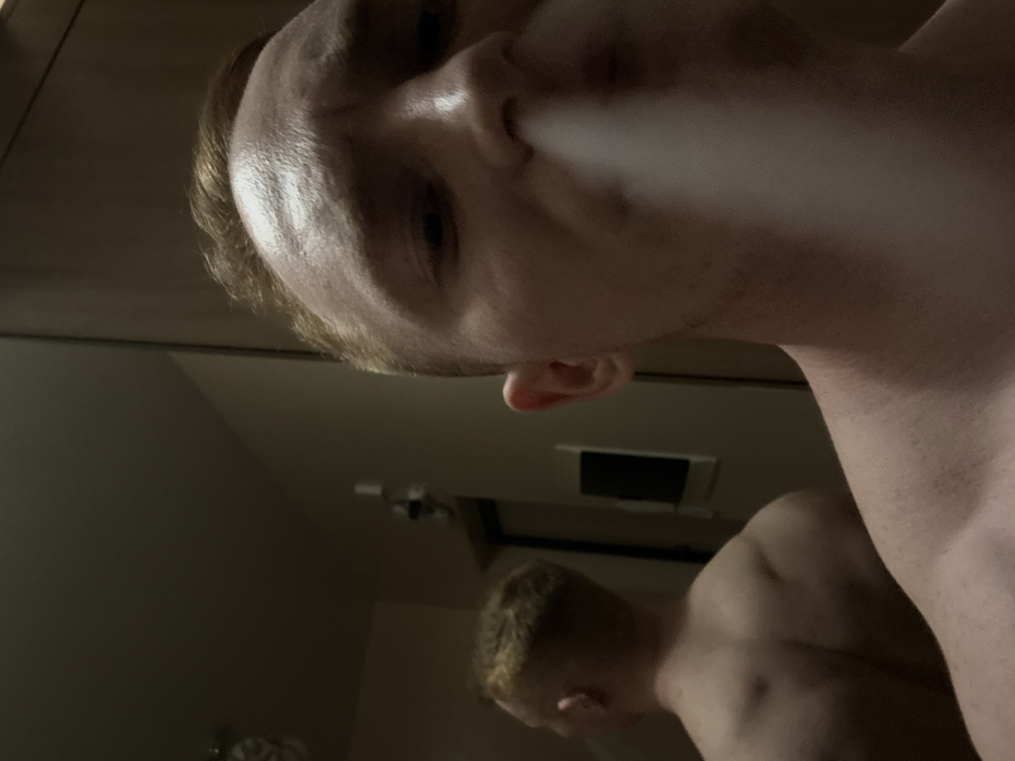The image presents a dim, close-up, and upward-angled shot of a young man, likely taken as a mirror selfie indoors. The man, with light-colored hair, occupies the upper part of the frame, exhaling a cloud of white smoke or vapor from his mouth, partially obscuring his lower face. He appears shirtless, with his bare shoulder visible on the right side of the image.
Below the main subject, a reflection of the man is visible in what appears to be a mirror. This reflection shows the back of his head, his ear, and his bare shoulders and upper back. The overall lighting is subdued and indirect, suggesting an indoor setting during the evening or in a dimly lit room. The background behind the man in the reflection is indistinct, appearing as dark, plain walls, with a rectangular object (possibly a vent or light fixture) and another blurry object visible. No specific architectural details or text are discernible to confirm the location context of Chaikovsky, Russia. The man's expression is neutral as he engages in the act of exhaling.