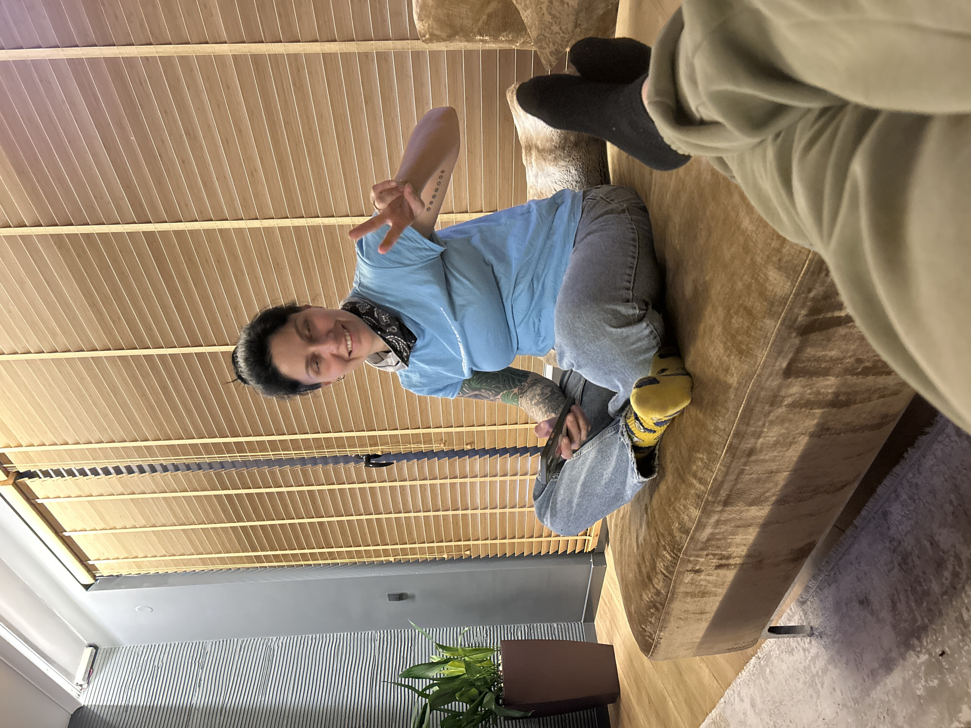 A person is sitting on a brown sofa, holding up a peace sign with their right hand. The person is wearing a blue t-shirt, gray jeans, and yellow patterned socks. The person is facing the camera and smiling. Another person is sitting next to them, wearing green pants and black socks. The background includes a wood-paneled ceiling, gray walls, and a potted plant. The location is most likely an apartment interior and there is no definitive proof of Sofia, Bulgaria.