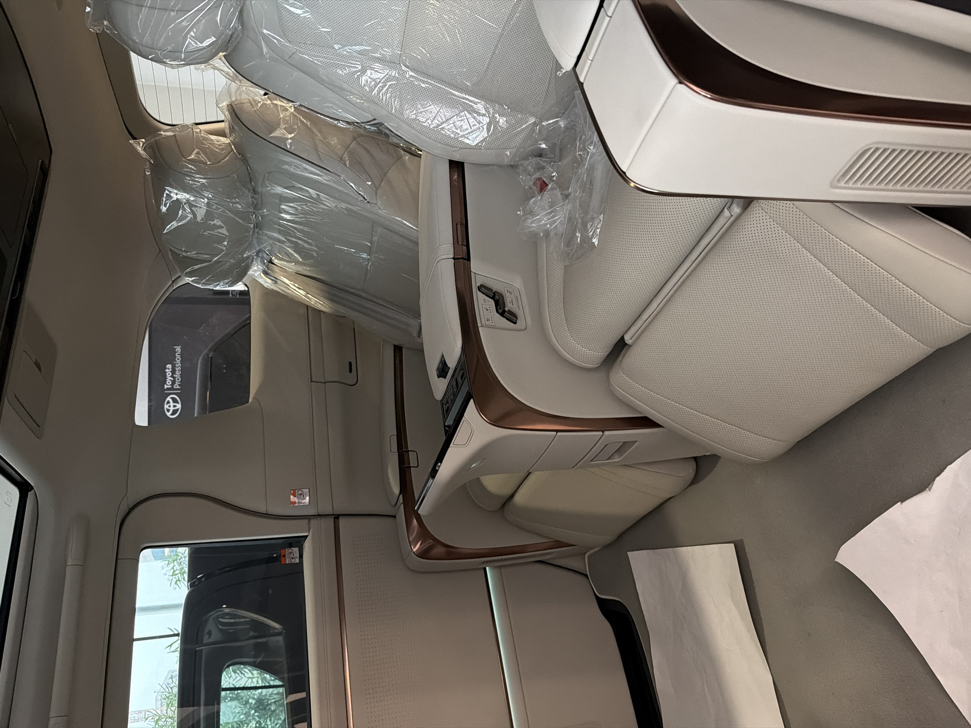 The image shows the interior of a luxury car, presumably a Toyota, given the "Toyota Professional" branding visible on a small display screen near a window. The seats are light-colored leather, covered in plastic wrap, suggesting the car is new or being transported/stored. The interior trim features bronze accents. The environment is clean and well-lit. The specific location within Podgorica, Montenegro, cannot be determined from the information in the image alone.
