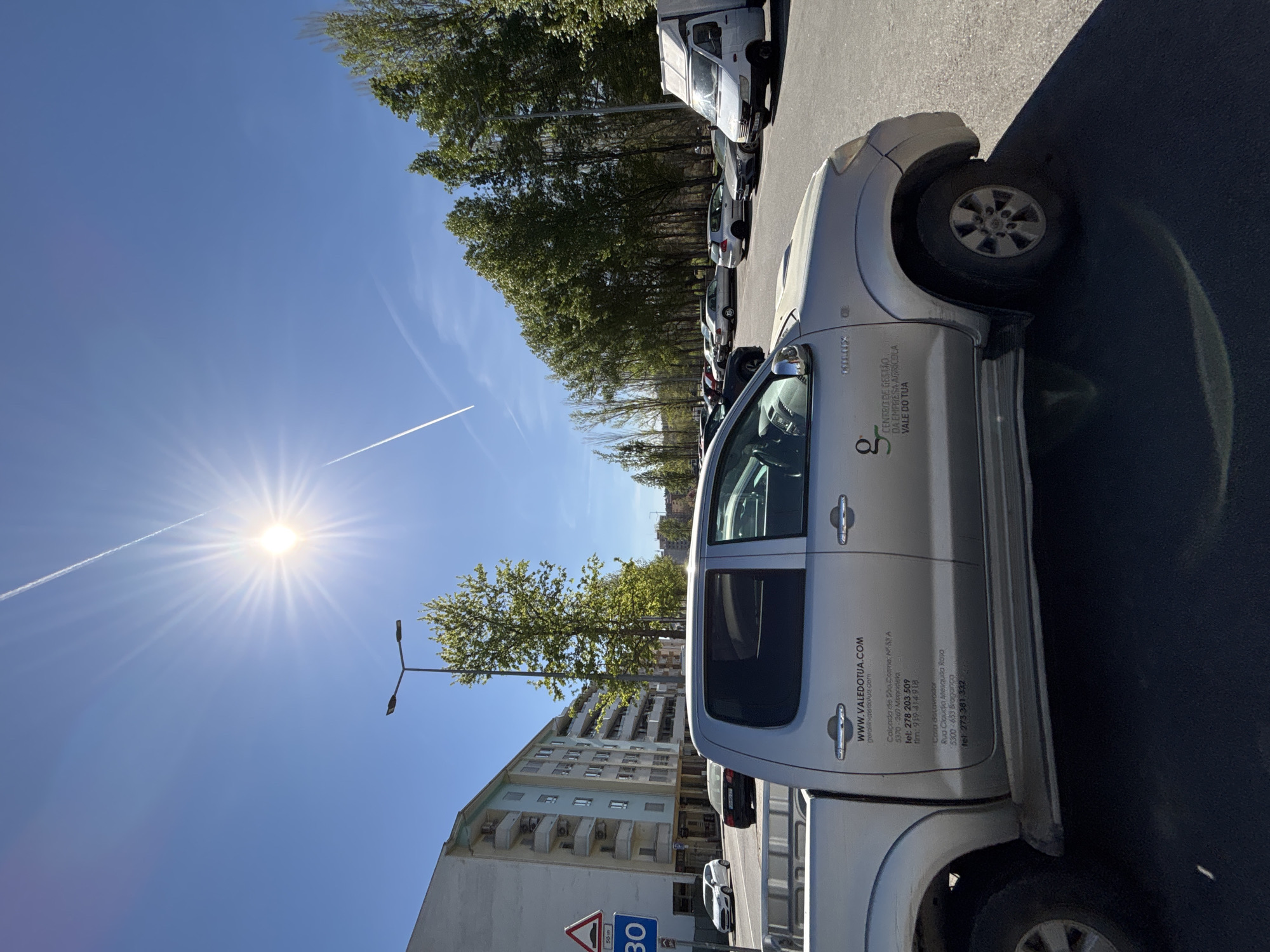 The image shows a silver Toyota Hilux pickup truck parked on an asphalt surface, likely a parking lot or street. The truck has the logo and contact information for "CENTRO DE GESTÃO DA EMPRESA AGRÍCOLA VALE DO TUA" printed on its side. The text also includes the website "WWW.VALEDOTUA.COM," a Mirandela phone number ("278 203 509"), and an address in Bragança ("Rua Claudio Mesquida Rosa"). In the background, there is a multi-story apartment building, street lights, trees, and other parked vehicles. A traffic sign indicating a speed limit (30) is visible. The sky is bright blue with the sun visible and a contrail from an airplane.