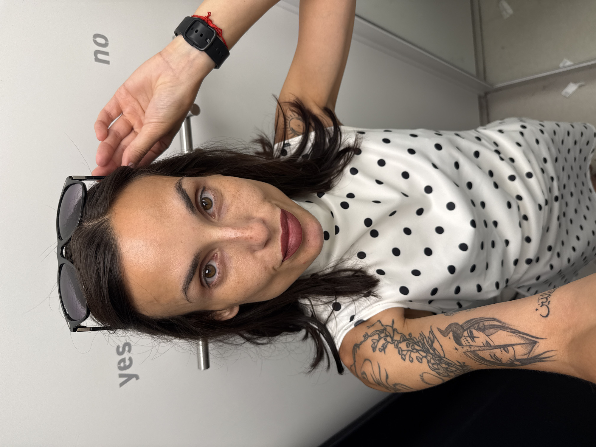 A woman with brown eyes and dark hair poses for a selfie in what appears to be a dressing room. She is wearing a white dress with black polka dots. She has a black digital watch on her wrist. There is a tattoo on her left arm. The wall behind her has the words "yes" and "no". The photograph was likely taken in a retail establishment, possibly in Pleven, Bulgaria.