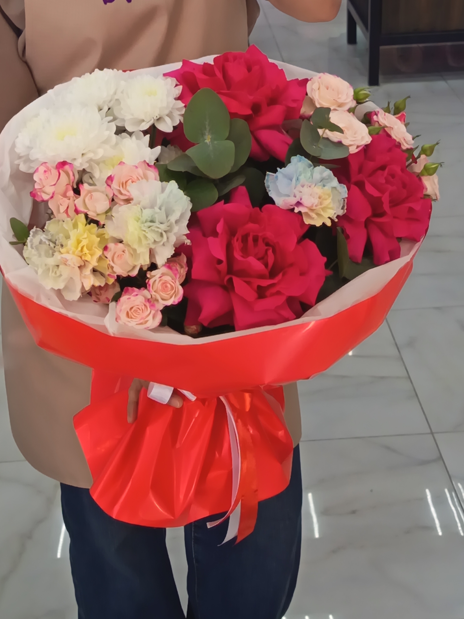 A person is holding a large bouquet of flowers wrapped in red paper with a white ribbon. The bouquet contains red roses, white chrysanthemums, and small pink and yellow flowers. The person is wearing a beige shirt and blue jeans. The background appears to be a tiled floor and a dark-colored piece of furniture. The location is unknown, and there is no indication in the image that the location is Nizhnevartovsk, Russia.