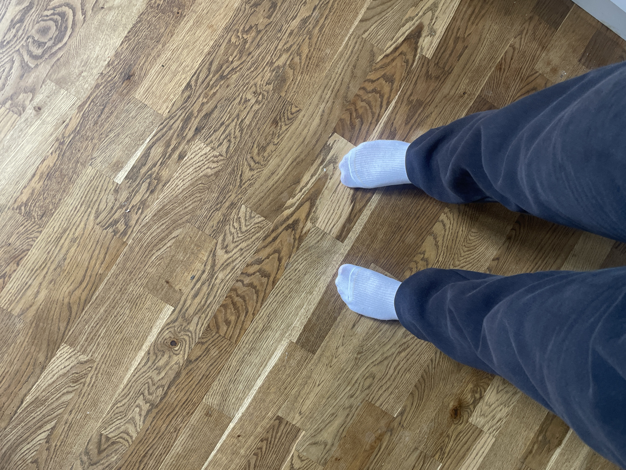 The image shows a person's legs and feet on a wooden floor. The person is wearing dark-colored sweatpants and white socks. The floor is made of light brown wood planks with a visible grain pattern. The photograph appears to be taken indoors. The image doesn't contain enough information to determine its location in the city of Maidstone, United Kingdom, or to describe any specific actions or interactions.
