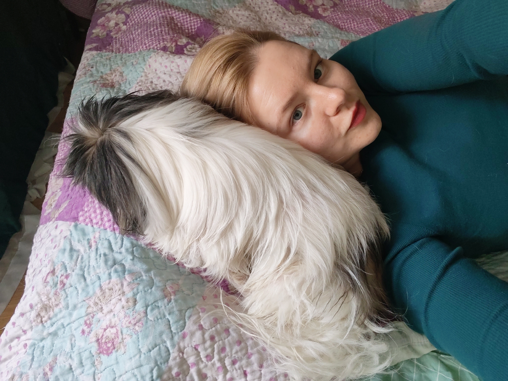 A fair-skinned woman with blonde hair and wearing a teal long-sleeved shirt lies on a bed with a patterned quilt, cradling a black and white Shih Tzu dog. The background appears to be indoors. There is no information to verify that the photo was taken in Izhevsk, Russia.