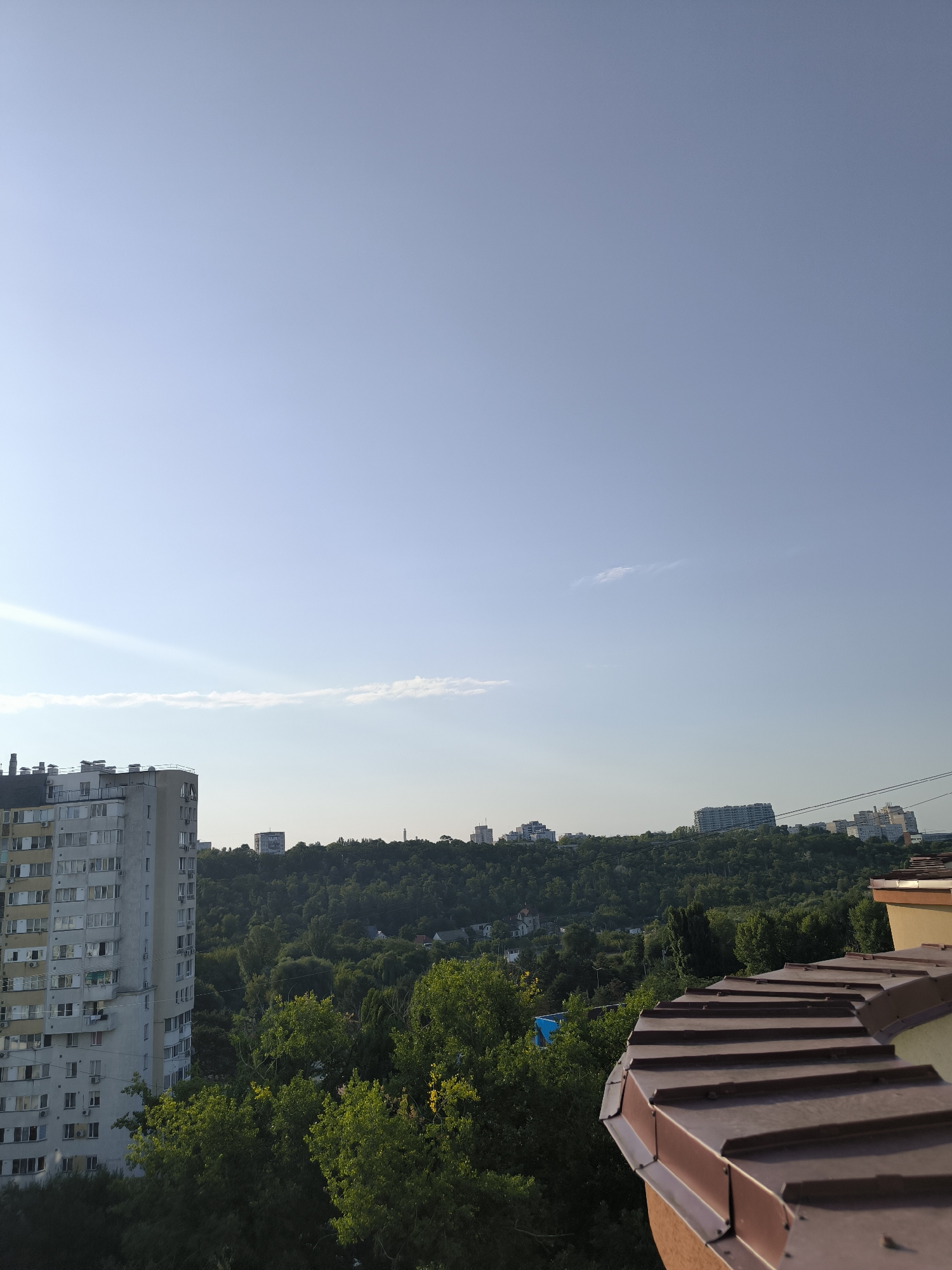 The image depicts an urban landscape with a clear blue sky above. In the foreground, a brown, tiled roof is visible, curving towards the right side of the frame. To the left, a tall, light-colored apartment building with numerous windows and balconies dominates the scene. Behind these structures, a dense, green forest covers rolling hills. Several other buildings, including apartment complexes and smaller houses, are scattered throughout the landscape, some visible on the hills and others peeking through the trees. Power lines are also present in the upper right corner of the image. The overall impression is of a city in Moldova, likely Chișinău, with a mix of residential buildings and natural greenery. The lighting suggests it is daytime, possibly late afternoon, given the angle of the sun casting shadows. There are no people or animals visible in the frame.