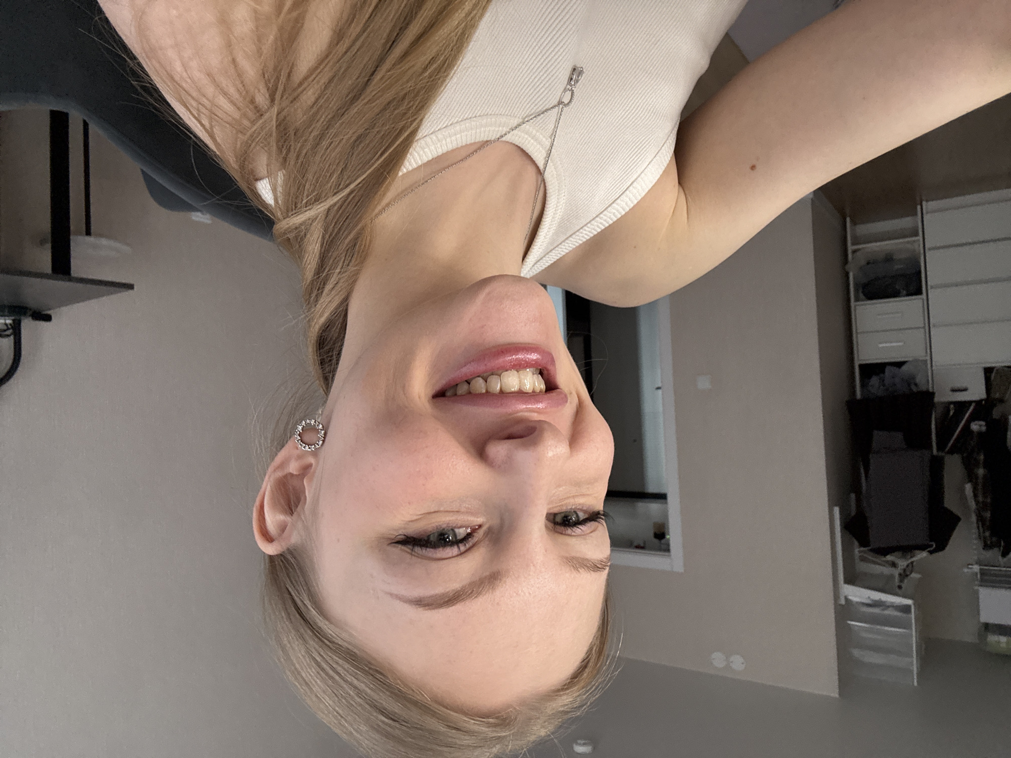 A young woman with long blonde hair, wearing a white tank top and circular earrings, smiles at the camera. She is indoors, with a chair visible to the left and a cabinet and window visible in the background. The image was taken in a residential area, likely within a home, in Perm, Russia.