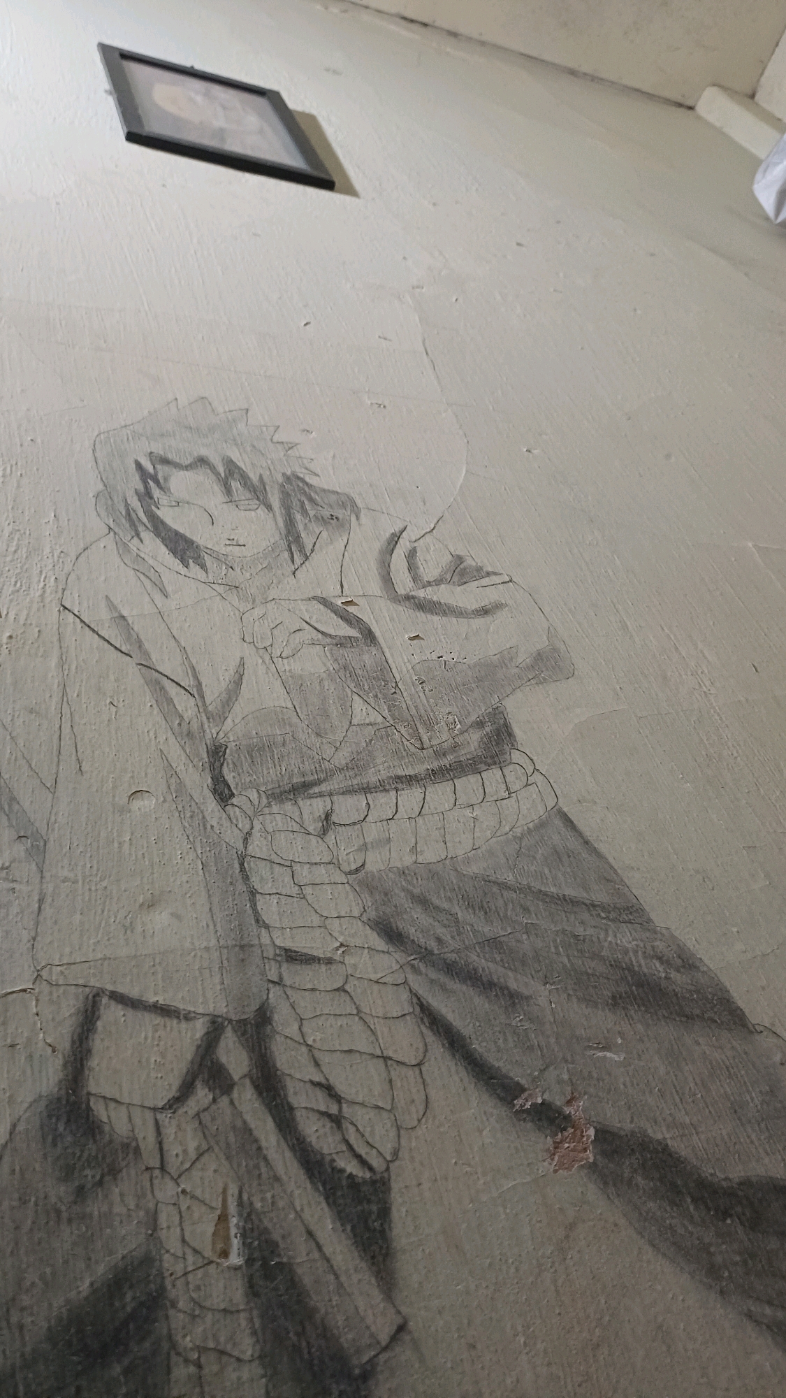 An unfinished pencil drawing of a male anime character, likely Sasuke Uchiha from Naruto, is depicted on a textured, off-white wall. The character is shown from approximately the waist up, with spiky black hair, a serious expression, and is wearing a dark robe with a banded sash around the waist. The drawing is rendered with varying degrees of shading, with some areas more detailed than others. The wall itself shows signs of age and wear, with cracks and peeling paint, particularly noticeable in the lower right portion of the image. A framed picture is hung on the wall in the upper left corner. The overall impression is of an artistic endeavor on a utilitarian surface in a nondescript indoor setting. The image does not provide information about the city of Wonokerso, Indonesia.