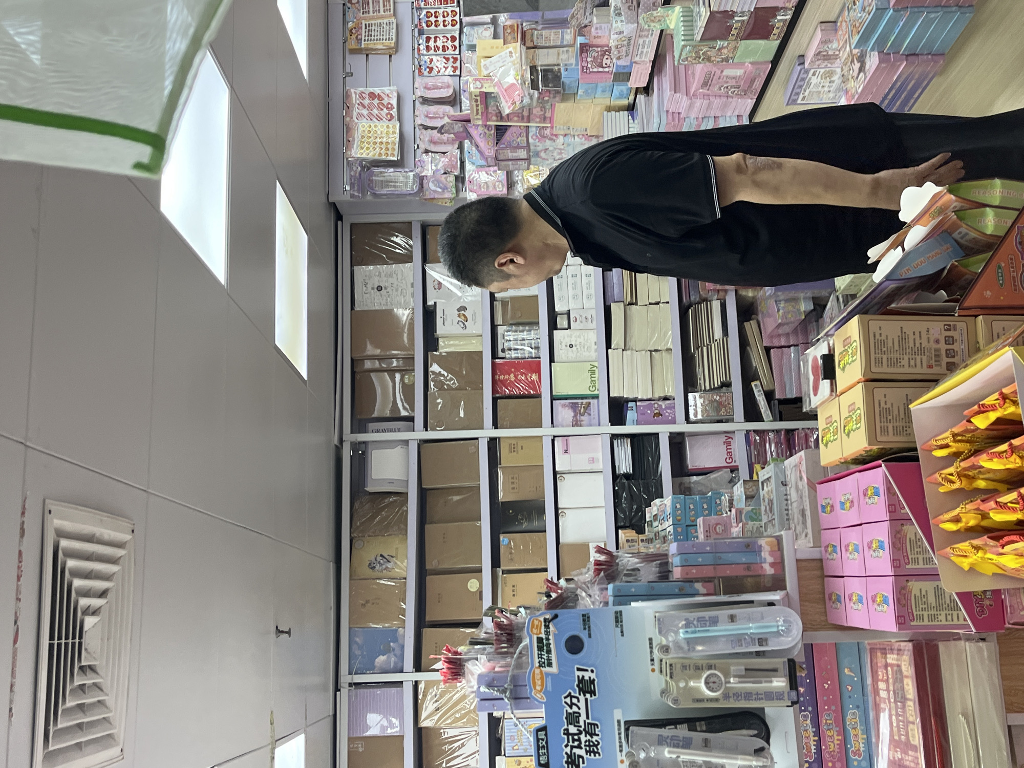 A man in a black shirt stands in a stationery store in Liwan District, China. The store is filled with shelves packed with notebooks, stationery sets, and various other paper products. The man is facing away from the camera, looking at the shelves. The lighting is bright, with fluorescent lights on the ceiling. The overall impression is of a well-stocked and organized store.
