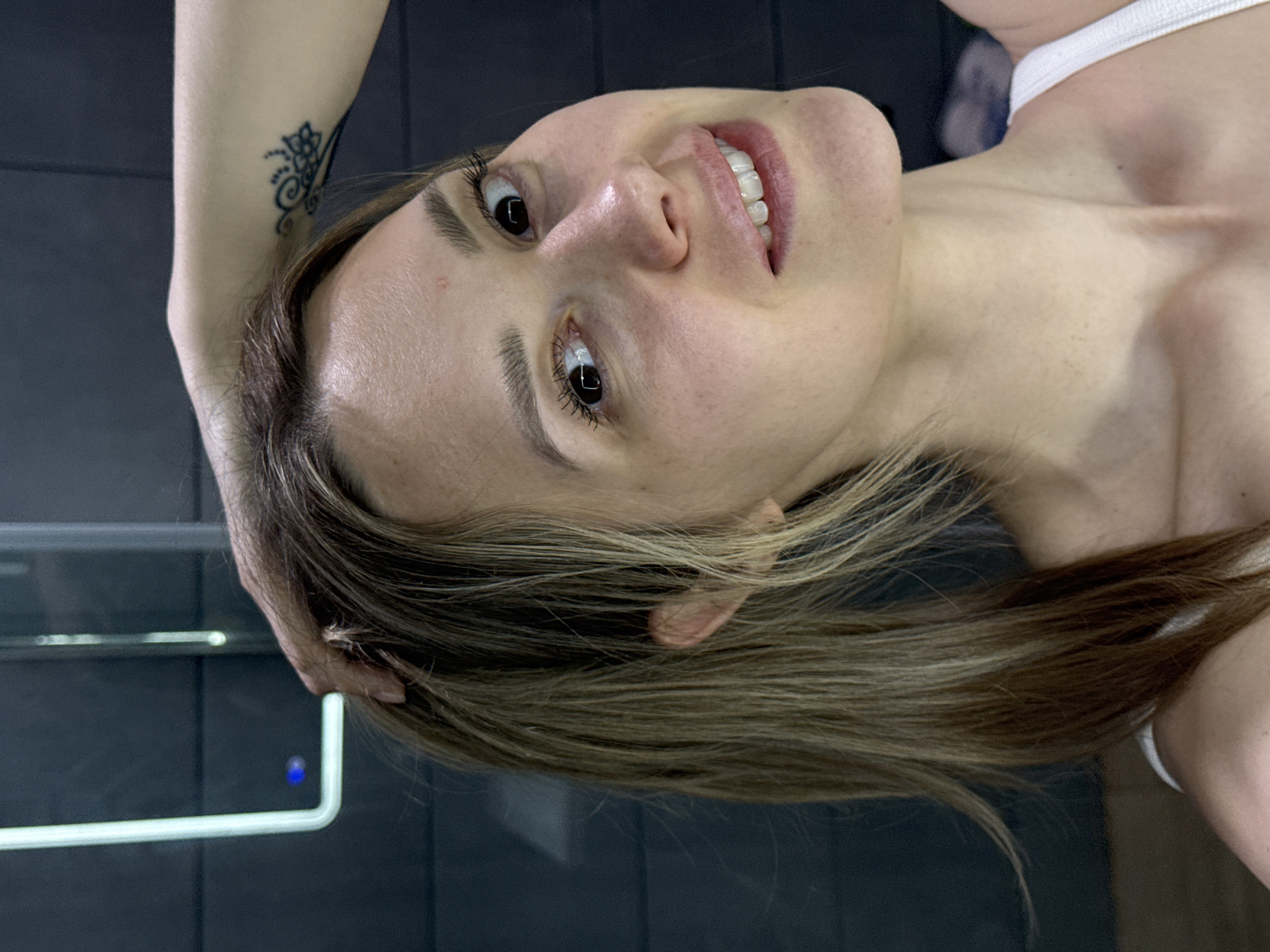 A medium-shot portrait of a light-skinned woman with long, blonde hair, likely taken in a bathroom. The woman is looking at the camera and smiling slightly. Her hair is partly behind her ear, and her hand is near her head, possibly holding her hair. She has a tattoo on her arm. The background appears to be a dark, tiled wall with a chrome fixture that includes a blue light indicator. There is insufficient information to verify the location as Chaikovsky, Russia.