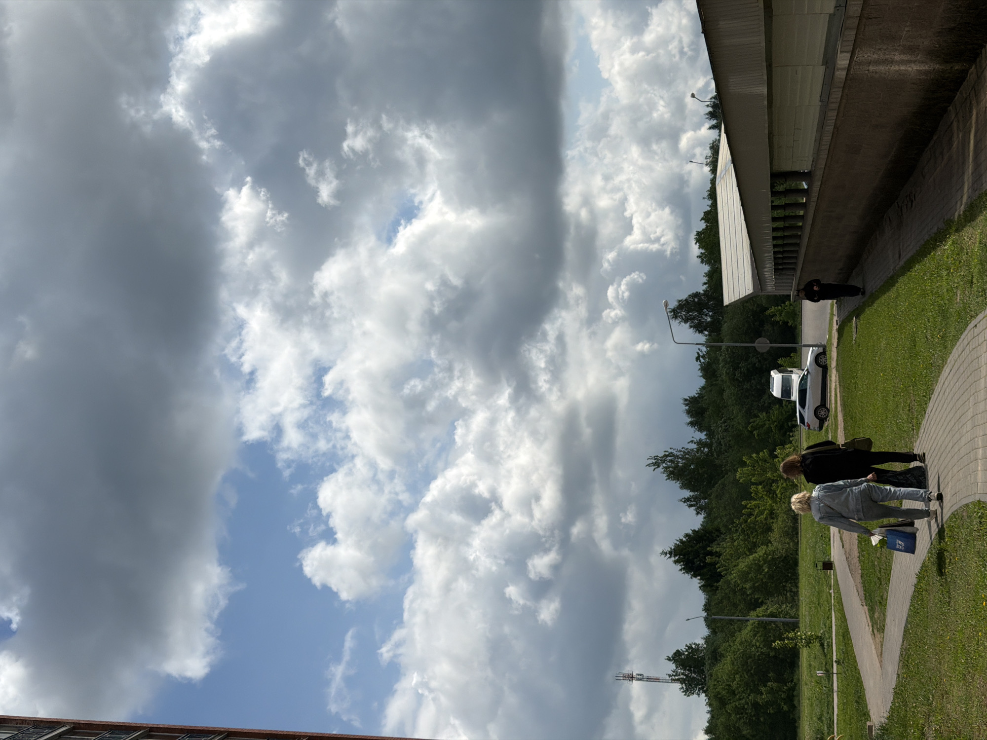 The image depicts an outdoor scene in an urban environment. Dominating the left side of the frame is a vast sky filled with heavy, billowing clouds, a mix of dark gray and bright white, contrasting with patches of blue sky.
The ground scene features a green, grassy area interspersed with trees. A paved walkway meanders through the area, occupied by two individuals walking together. The closer person wears a gray outfit and carries a blue bag. The other person is dressed in black. Nearby, a white vehicle is parked beside the walkway. In the background, there's a raised structure, possibly a bridge or elevated building, with a gray wall and green overhanging features. A figure stands beneath the structure. A lamppost is also visible in the scene.