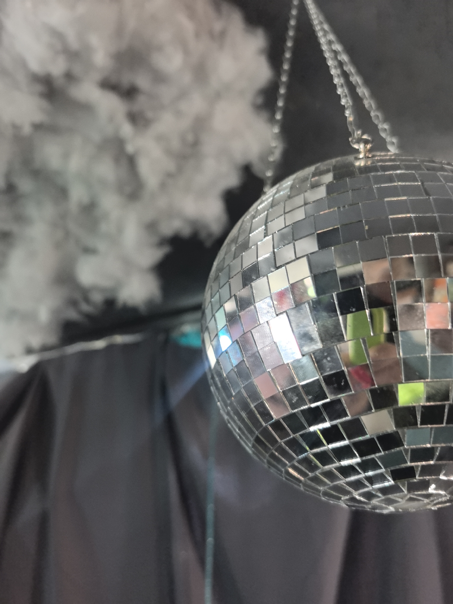 A disco ball hangs from a chain, suspended in front of a dark, draped background and wisps of white smoke or fog. The disco ball is made of numerous small mirrored squares that reflect the surrounding environment, showing flashes of color including green, pink, and blue. The smoke is dense in the upper left portion of the image and thins out towards the right. The scene appears to be indoors, possibly in a venue or event space, but its specific location within Noviy, Russia, cannot be determined from the image. There are no people visible in the image.