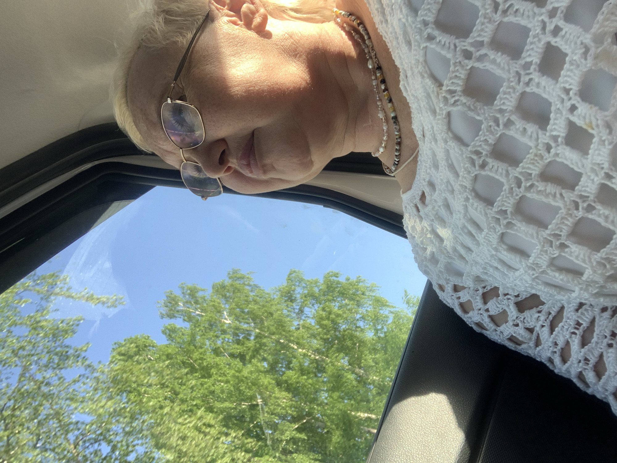 The image depicts a middle-aged Caucasian woman with short blonde hair and glasses looking out of a car window. She is wearing a white crocheted garment and a beaded necklace. The view through the window shows a clear blue sky and green trees, likely taken during the daytime. Considering the city specified, Chaikovsky, Russia, the environment suggests a temperate climate with deciduous trees. The angle of the shot indicates the photograph was likely taken from within the car, looking slightly upwards at the woman.