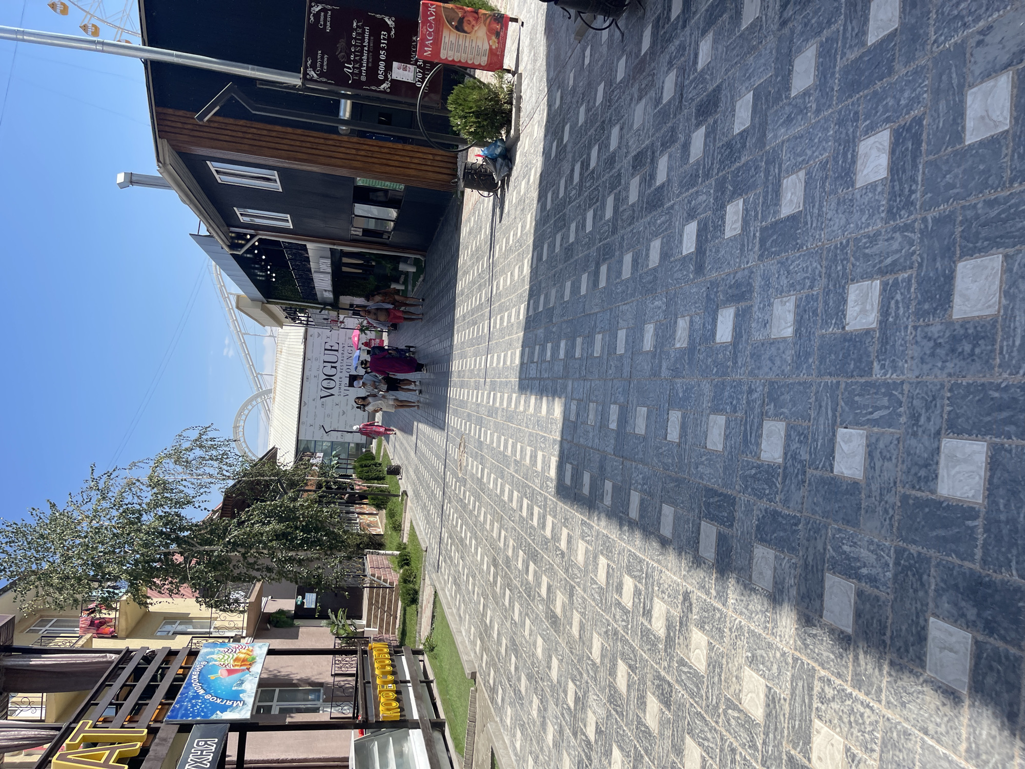 The image captures a scene on a paved walkway in Bosteri, Kyrgyzstan, likely a tourist area given the presence of a Ferris wheel in the background and shops lining the street. The walkway is paved with a repeating pattern of dark grey and light grey square tiles, with a prominent shadow cast across it from the left.
On the left side of the walkway, a building with balconies and a sign reading "КOФЕ С СОБОИ" (Coffee to go) is visible. Further down, a sign for "VOGUE SUMMER RESTAURANT VIP LOUNGE" is displayed. Several individuals are walking along the walkway, some appearing to be tourists.
On the right side, a dark-colored building features a sign for a massage parlor with the text "Массаж" (Massage) and a phone number. A red advertising stand for massage services is placed on the pavement. In the distance, a large white roller coaster-like structure and a Ferris wheel are visible against a clear blue sky. The environment appears to be an outdoor, daytime setting, with greenery interspersed along the walkway.
