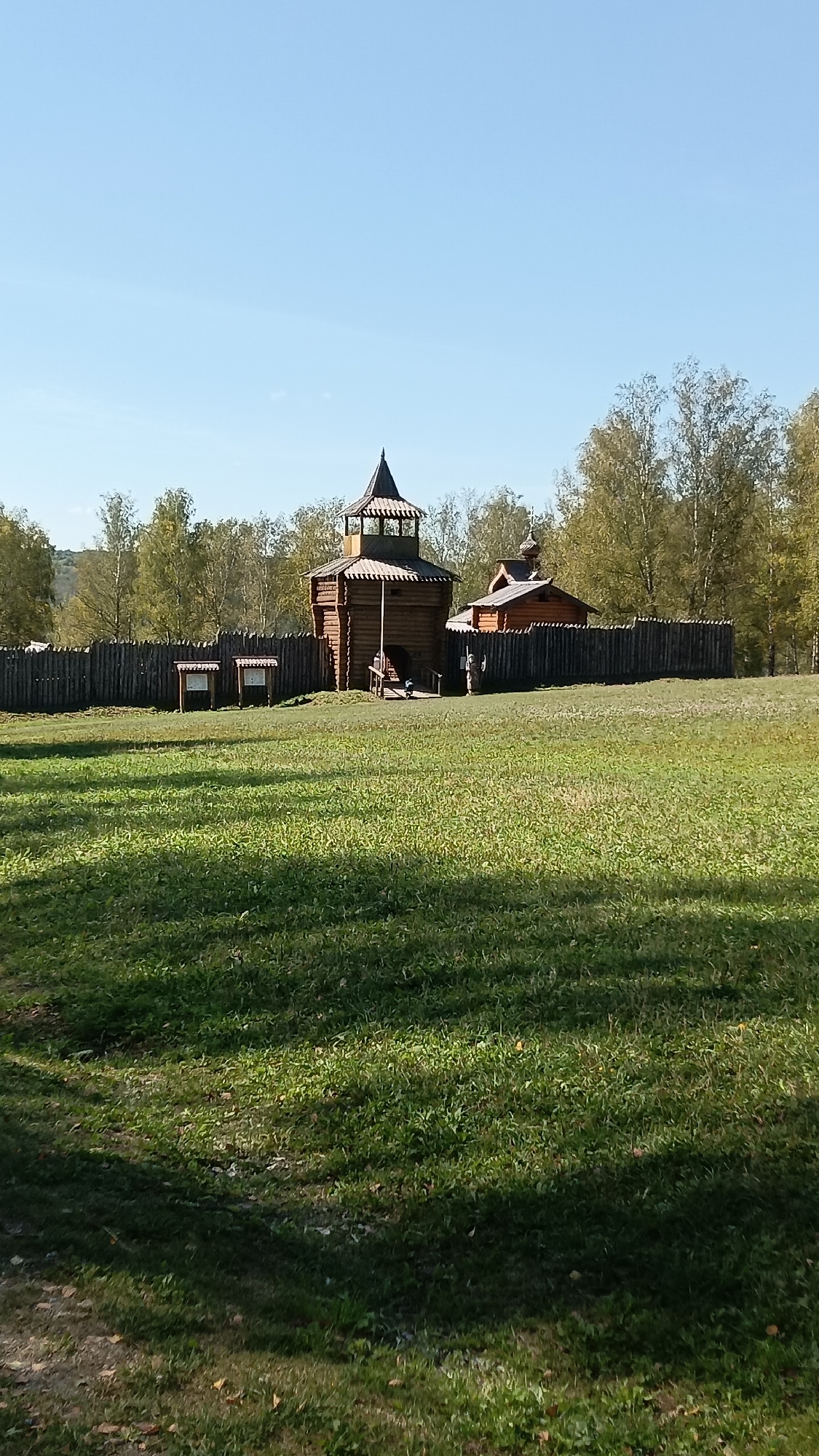 The image shows a wooden fortress with a tower and a small wooden church. The fortress is surrounded by a wooden fence. In front of the fortress, there is a green grassy field. Behind the fortress, there are trees and a clear blue sky. The scene appears to be an historical reconstruction or a park. There are no people visible in the image. The lighting suggests it is daytime. The scene is located in a rural or park-like setting.